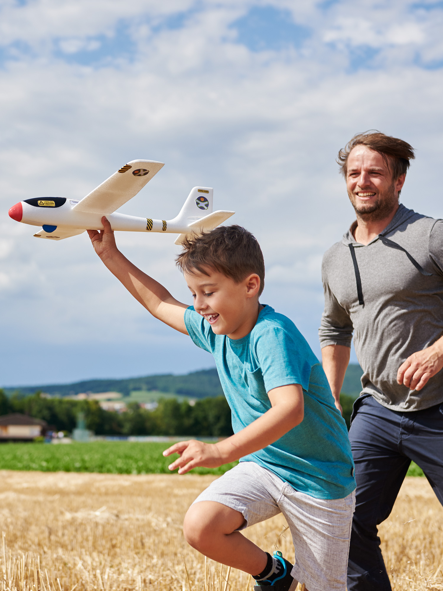 Ein Junge rennt mit einem Spielzeugflugzeug, gefolgt von einem Mann. Sie sind auf einem Feld mit blauem Himmel.