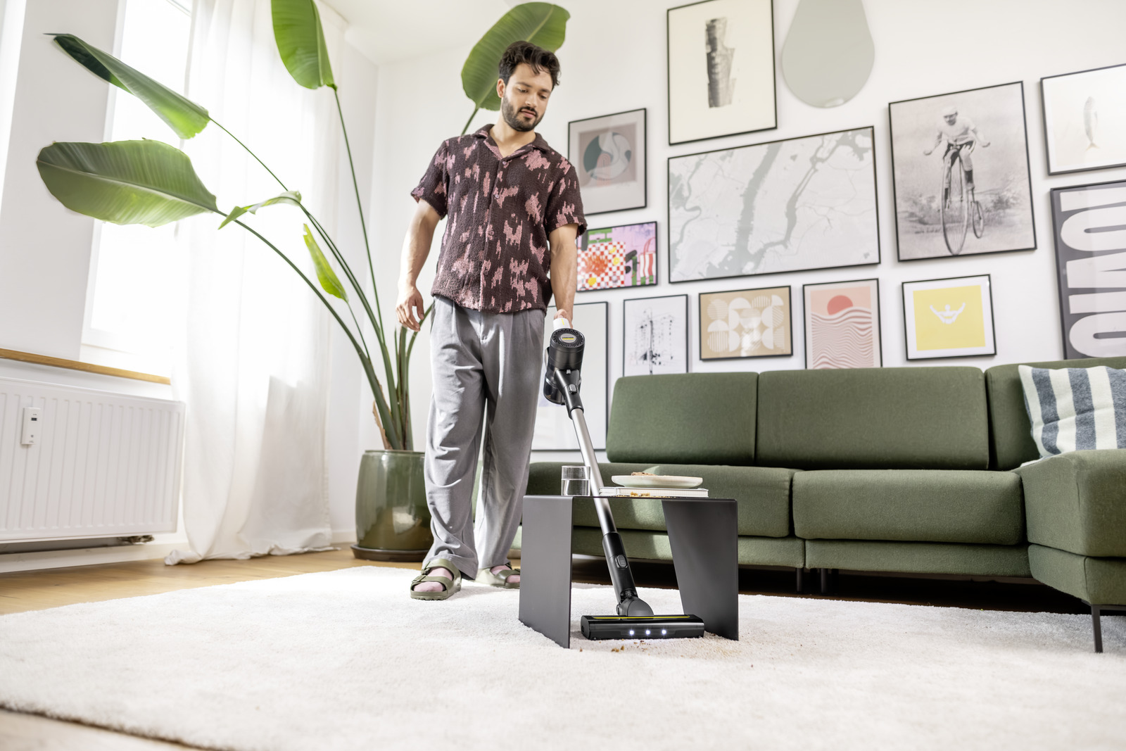 Mężczyzna odkurza pokój z białym dywanem, zieloną sofą i obrazami na ścianie. Duża roślina w rogu.