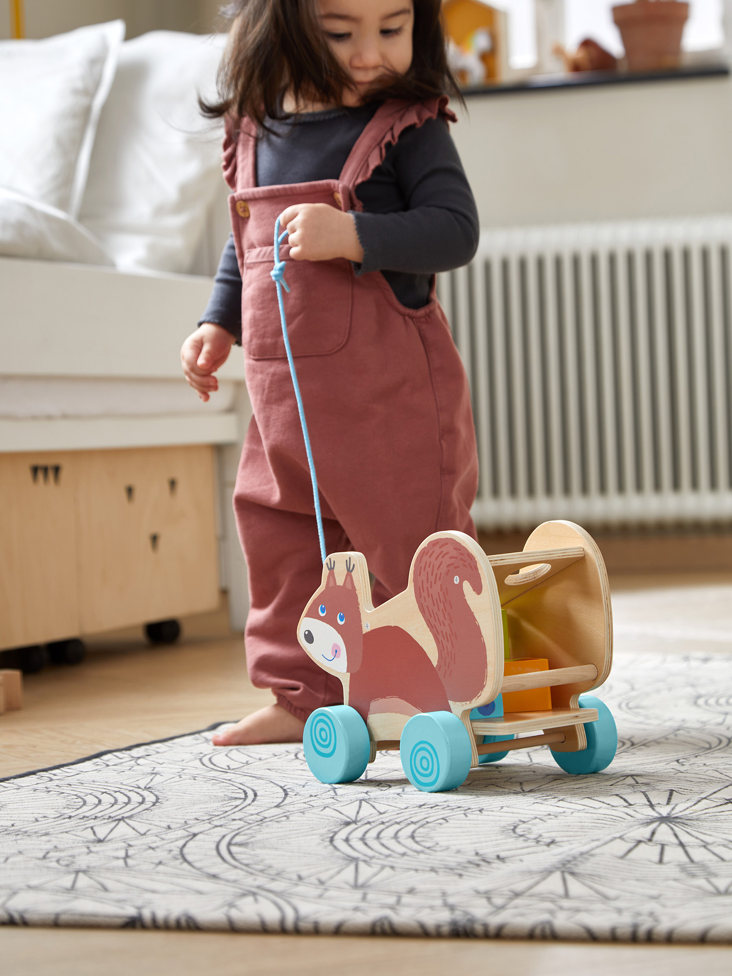 Ein Kleinkind in Overalls zieht einen eichhörnchenförmigen Spielzeugwagen auf einem gemusterten Teppich. Der Wagen hat blaue Räder, und der Hintergrund ist ein Zimmer.