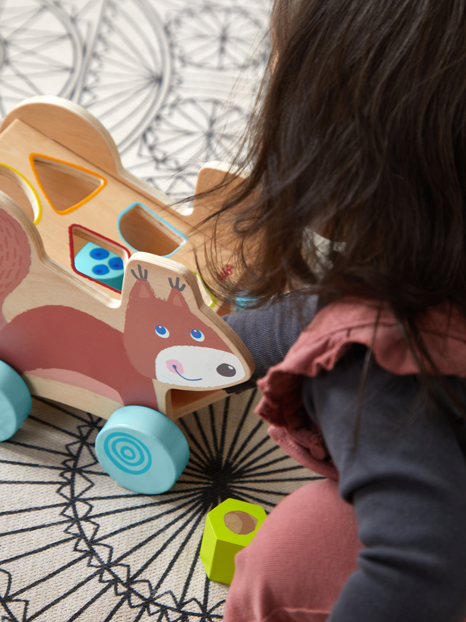 Ein Kind interagiert mit einem hölzernen Eichhörnchen-Spielzeugauto. Das Spielzeug hat blaue Räder. Das Kind trägt ein rosa Oberteil.