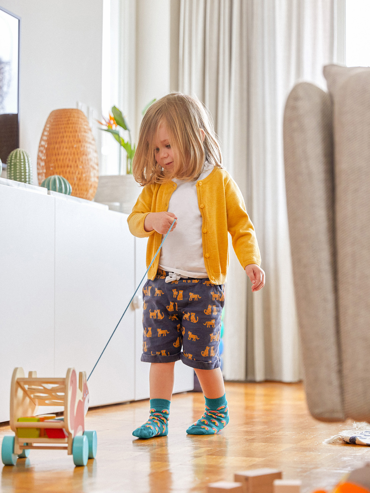 Ein junges Kind in gelber Strickjacke zieht einen hölzernen Spielzeugwagen. Das Kind trägt Shorts mit Katzenprints und blaue Socken.