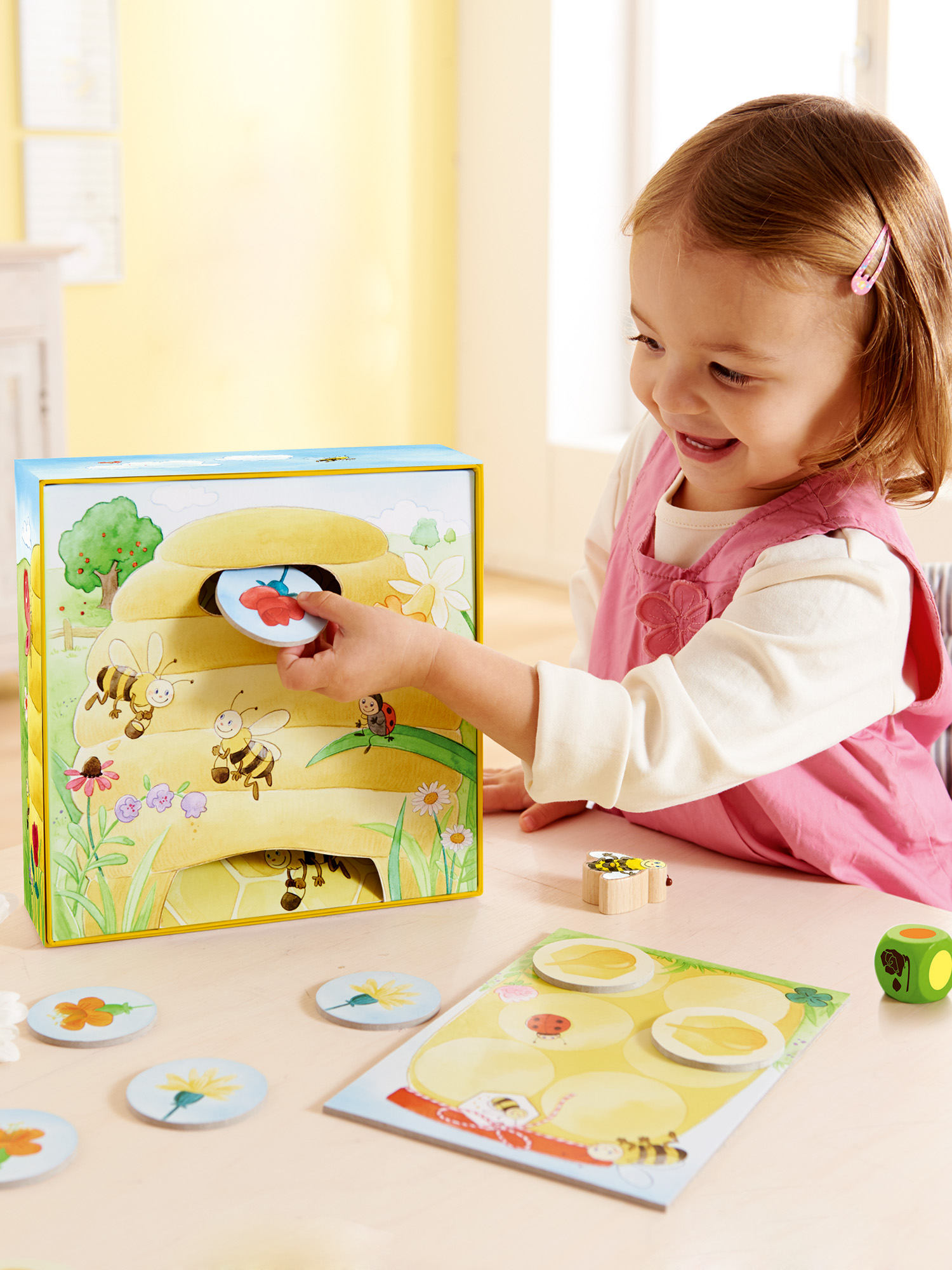 Ein Mädchen spielt ein Bienen-Zuordnungsspiel. Sie steckt eine Scheibe in eine bienenstockförmige Box. Spielsteine und ein Würfel liegen auf dem Tisch.