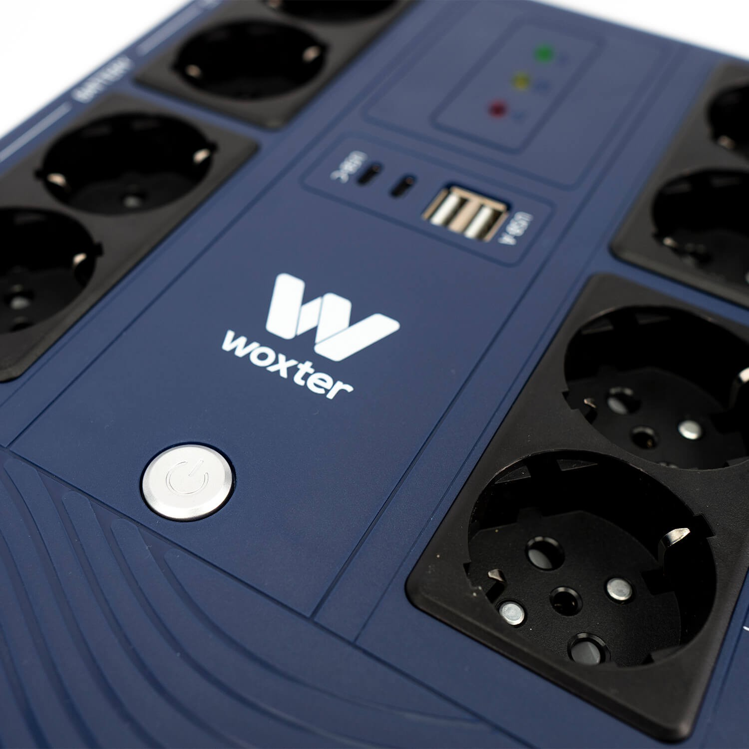 Una regleta azul con enchufes negros, un botón de encendido blanco y el logo "woxter". También se ven un puerto USB y luces indicadoras.