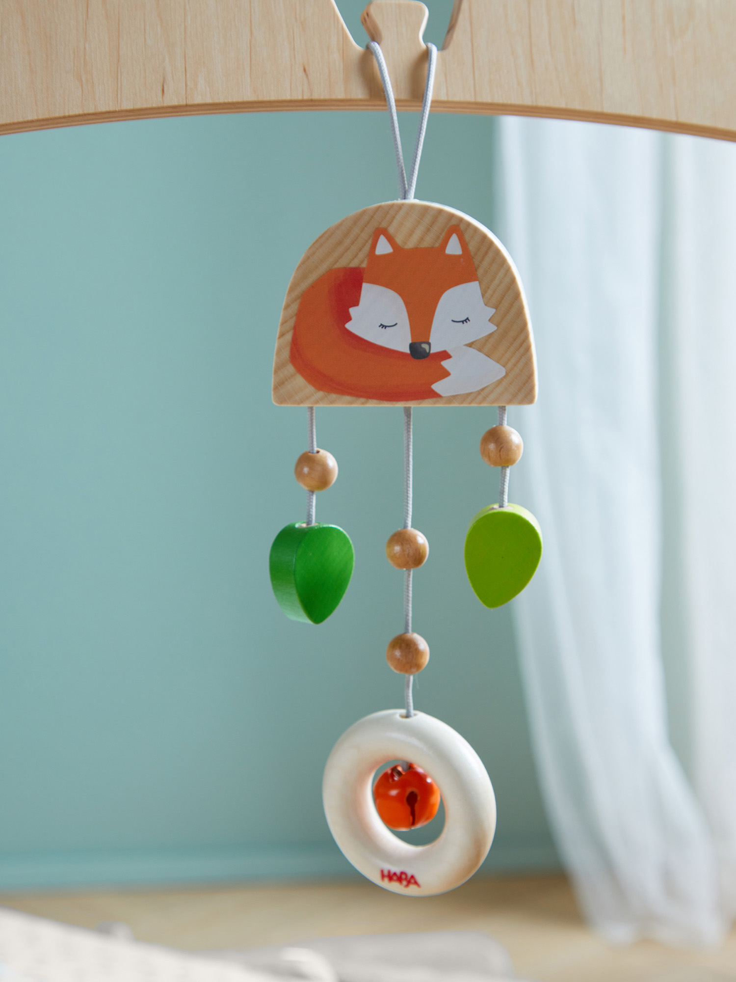 Holz-Babymobile mit Fuchsmotiv, grünen Blättern, Holzkugeln und einer Glocke. Der Hintergrund ist hellblau und ein weißer Vorhang.
