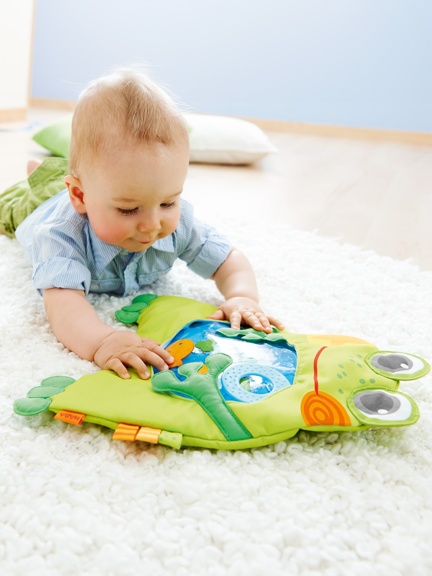 Ein Baby liegt auf einem weißen Teppich und betrachtet ein grünes Froschspielzeug. Das Spielzeug hat orangefarbene und blaue Details.
