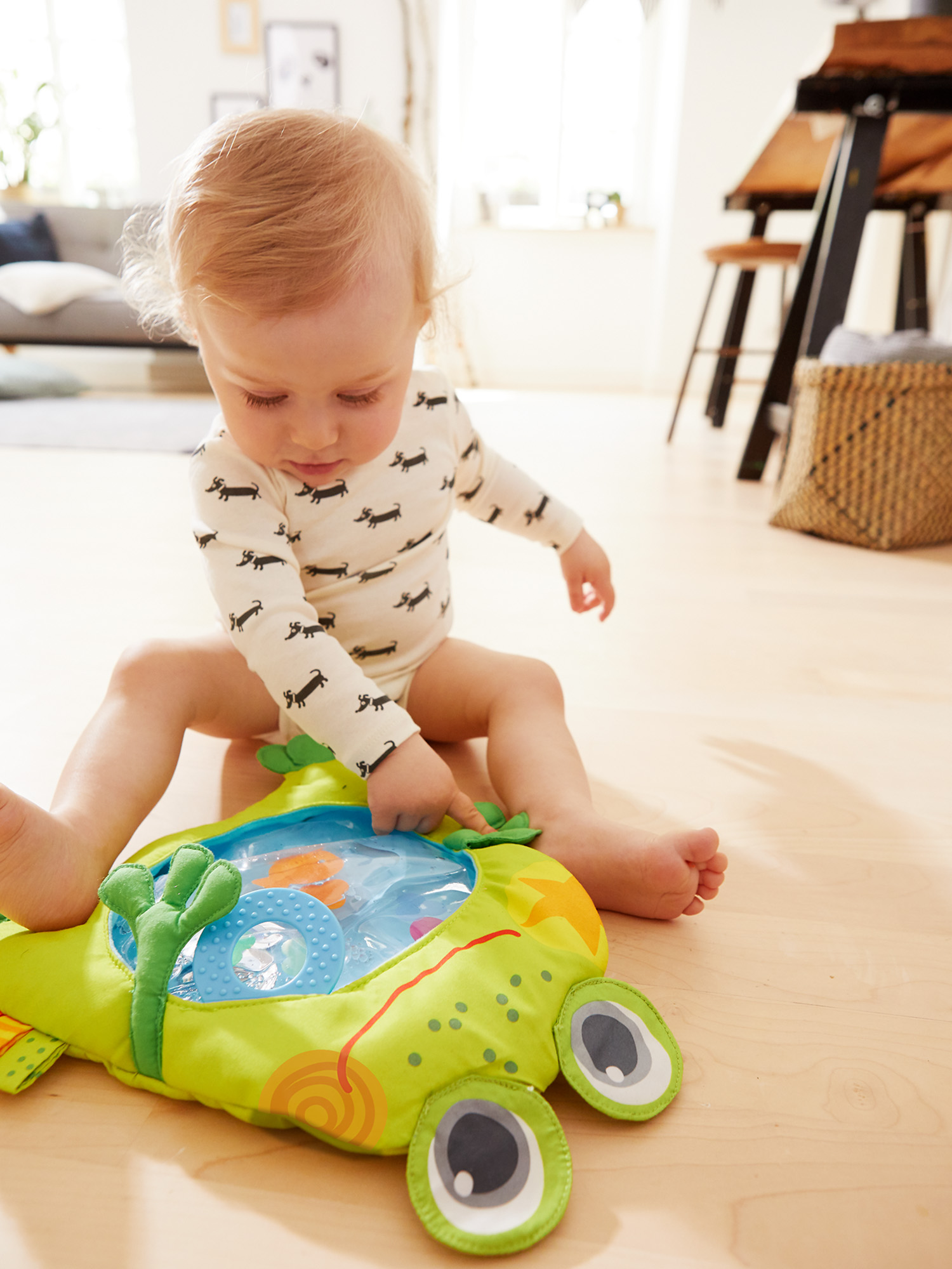 Ein Baby sitzt auf dem Boden und betrachtet ein grünes Froschspielzeug. Das Spielzeug hat einen blauen Wasserteil mit schwimmenden Gegenständen. Das Baby trägt einen Onesie mit Hundeaufdruck.