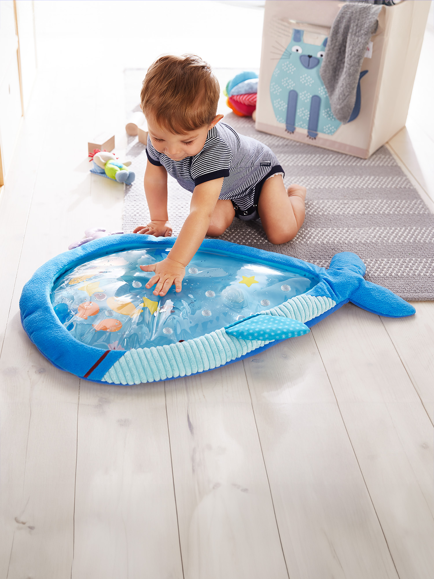 Ein Baby berührt eine walförmige Wassermatte. Die Matte liegt auf einem Holzboden. Spielzeug und eine Aufbewahrungsbox sind im Hintergrund.