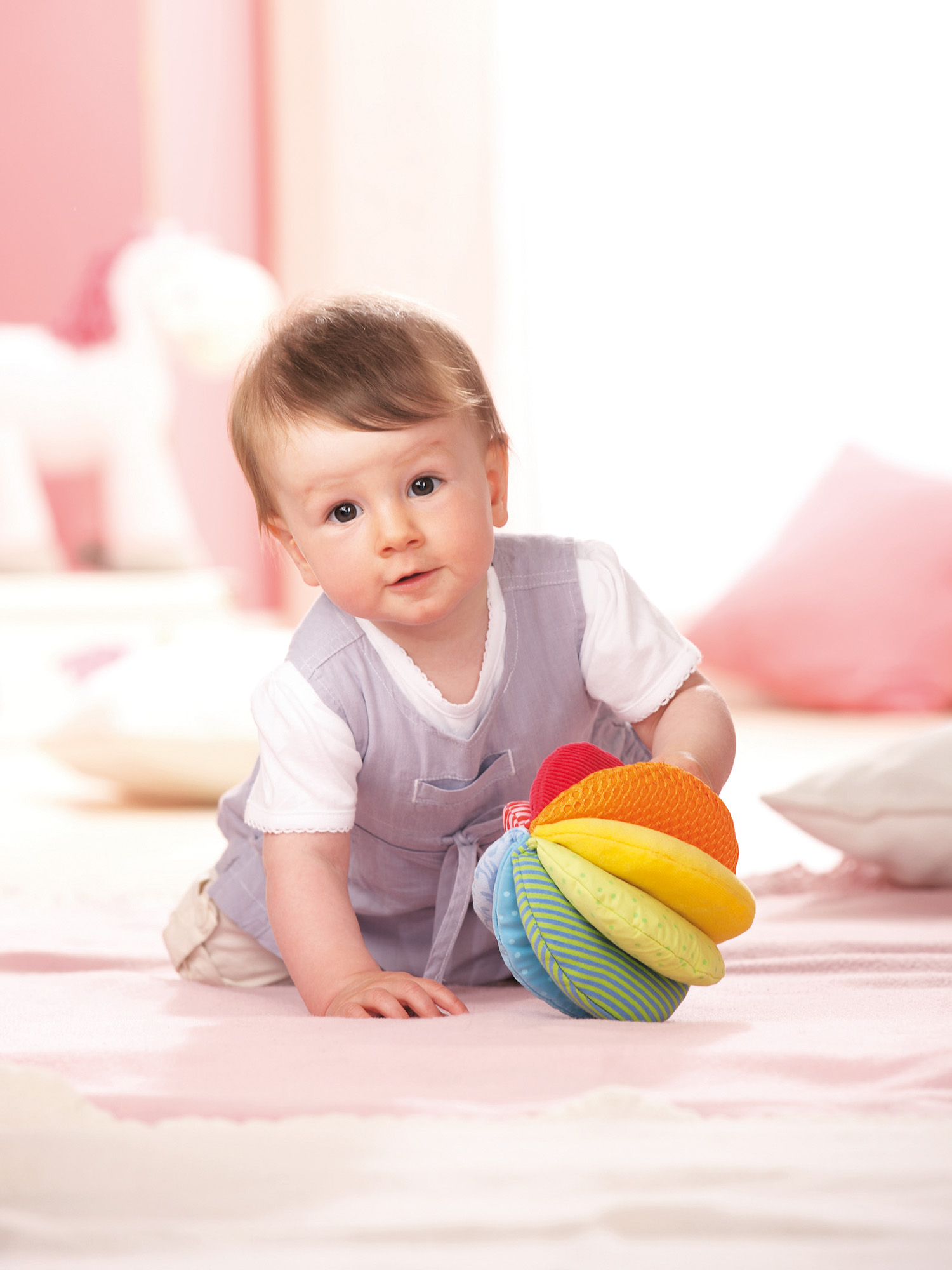 Ein Baby krabbelt auf einer rosa Oberfläche und hält ein buntes Spielzeug. Kuscheltiere und Kissen im Hintergrund deuten auf ein Kinderzimmer hin.