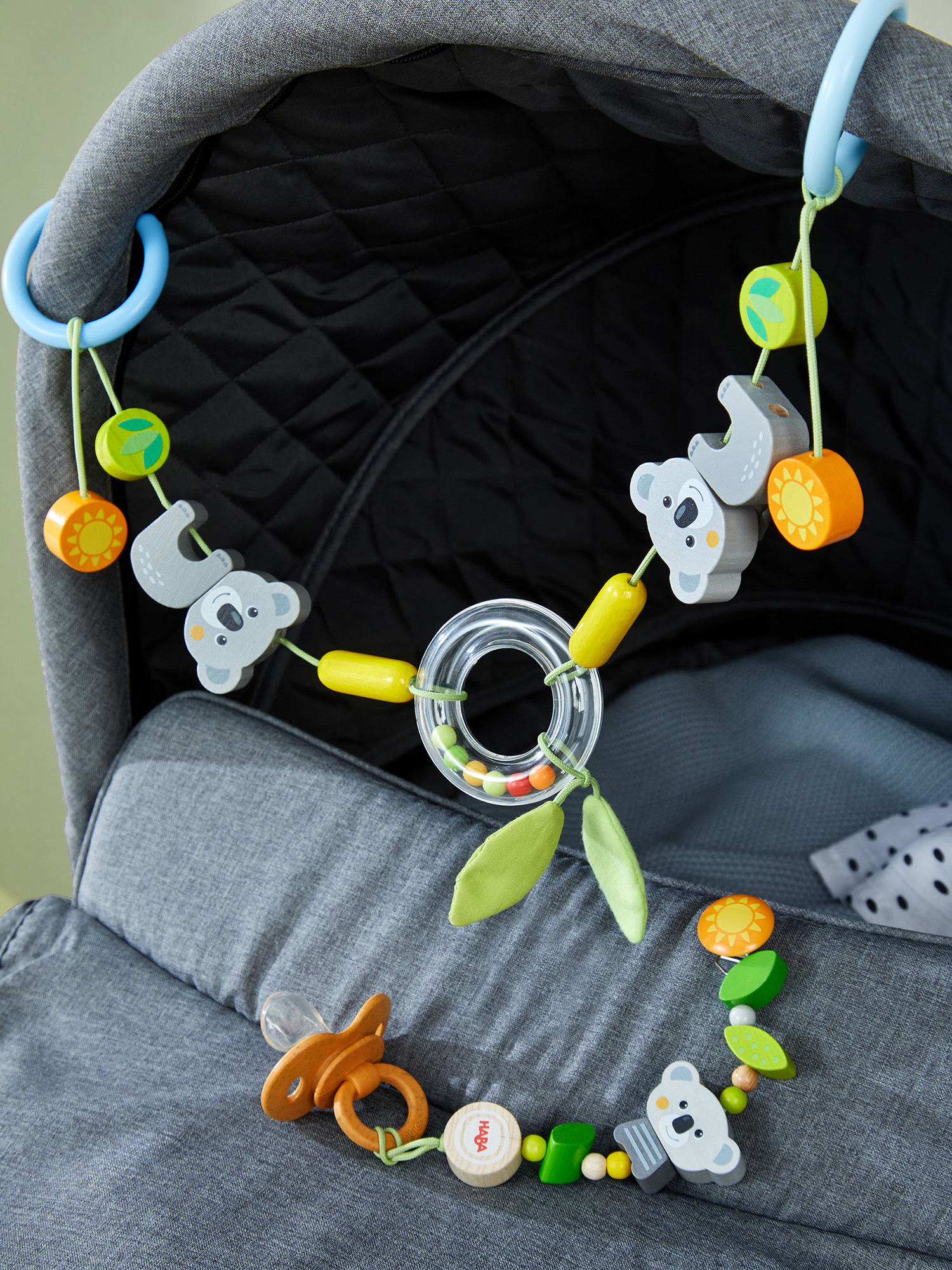 Ein Kinderwagen mit einem Holzspielzeug-Mobile mit Koalas, Sonnen und Blättern. Darunter liegt ein Schnuller. Grauer und schwarzer Stoff.