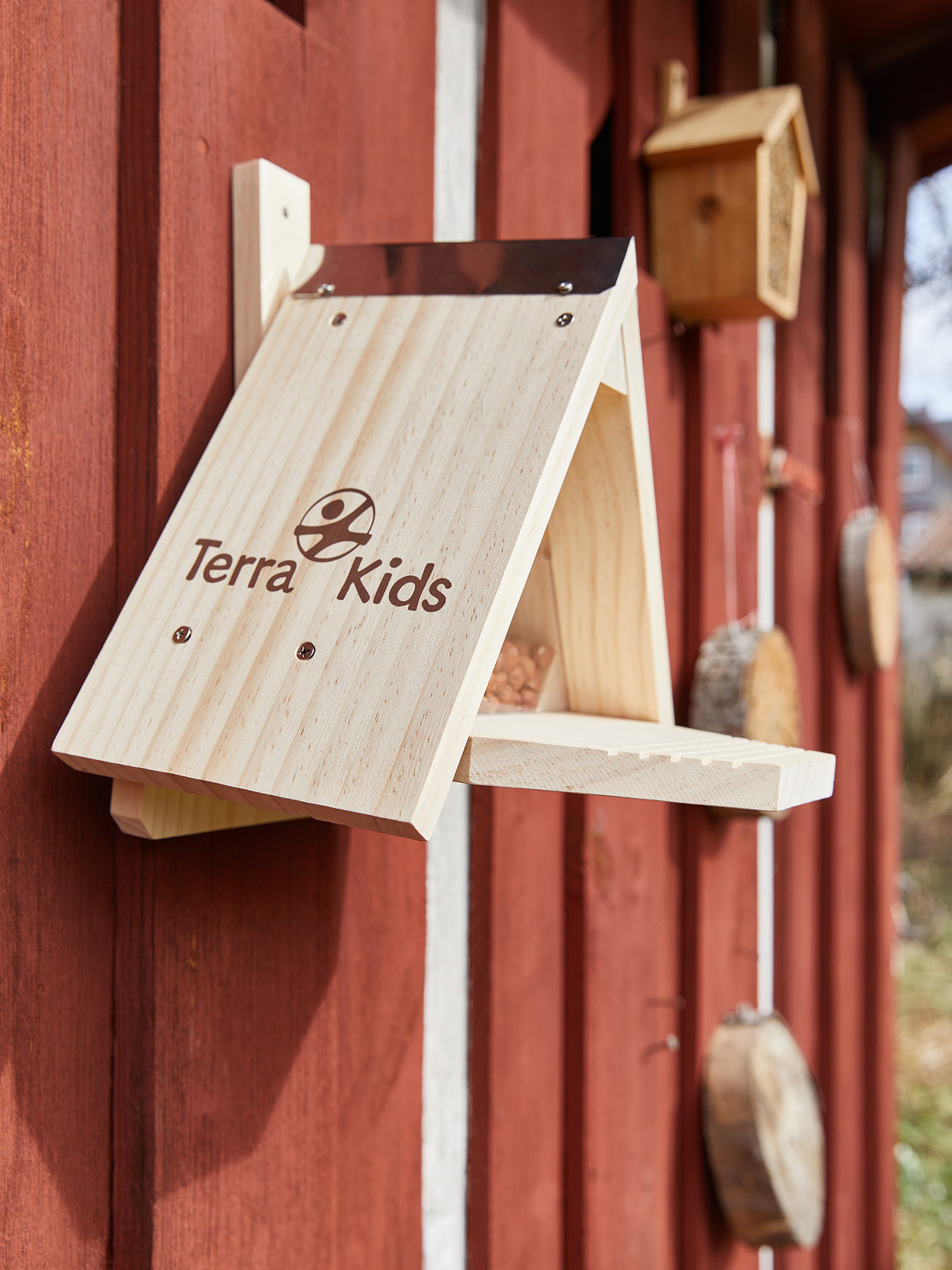 Ein hölzerner Vogelfutterautomat mit dem Logo 'Terra Kids' ist an einer roten Holzwand befestigt. Ein kleines Vogelhaus und Holzscheiben hängen in der Nähe.