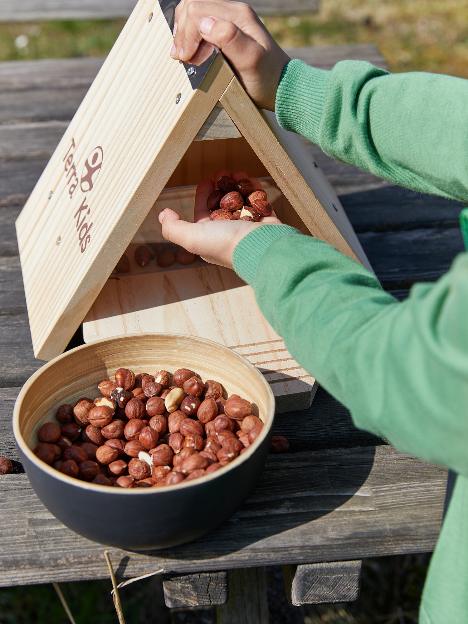 Eine Person in Grün füllt ein Vogelhaus aus Holz mit Haselnüssen. Eine Schüssel mit Haselnüssen steht auf einem Holztisch.