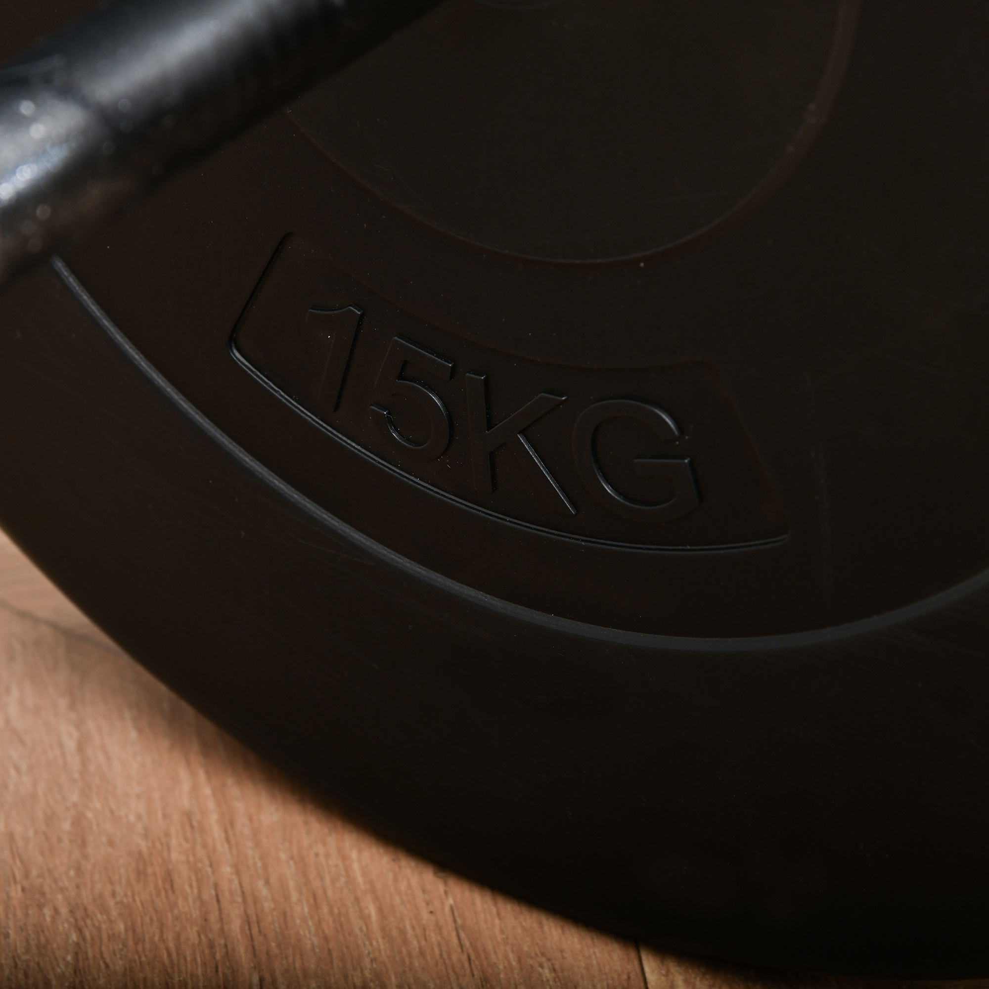Plato de pesas negro con texto blanco en relieve '15KG'. Una barra es visible. Suelo de madera al fondo.