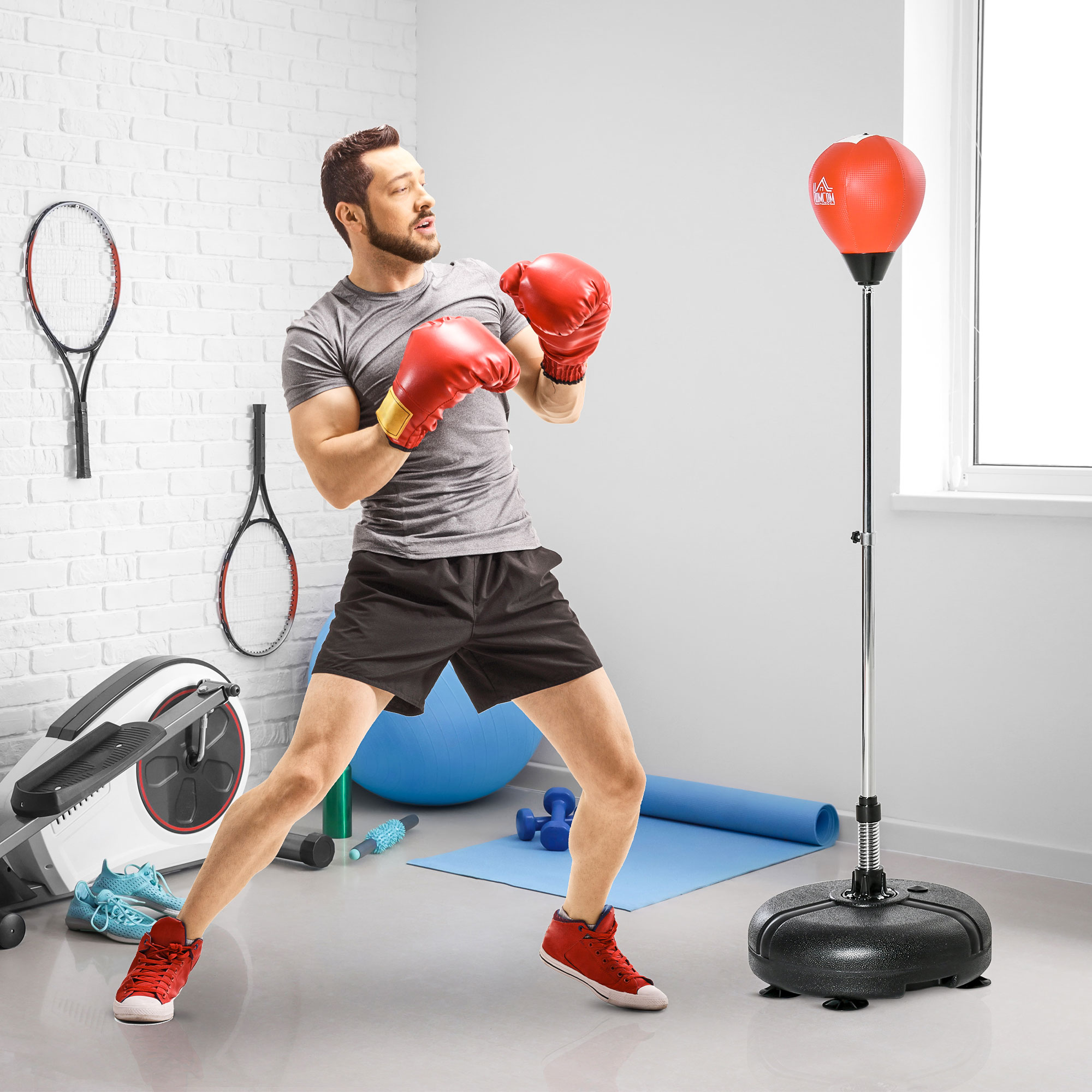 Hombre en ropa deportiva boxea un saco de boxeo rojo. Raquetas de tenis colgadas en la pared. Equipos de gimnasio y una esterilla azul al fondo.