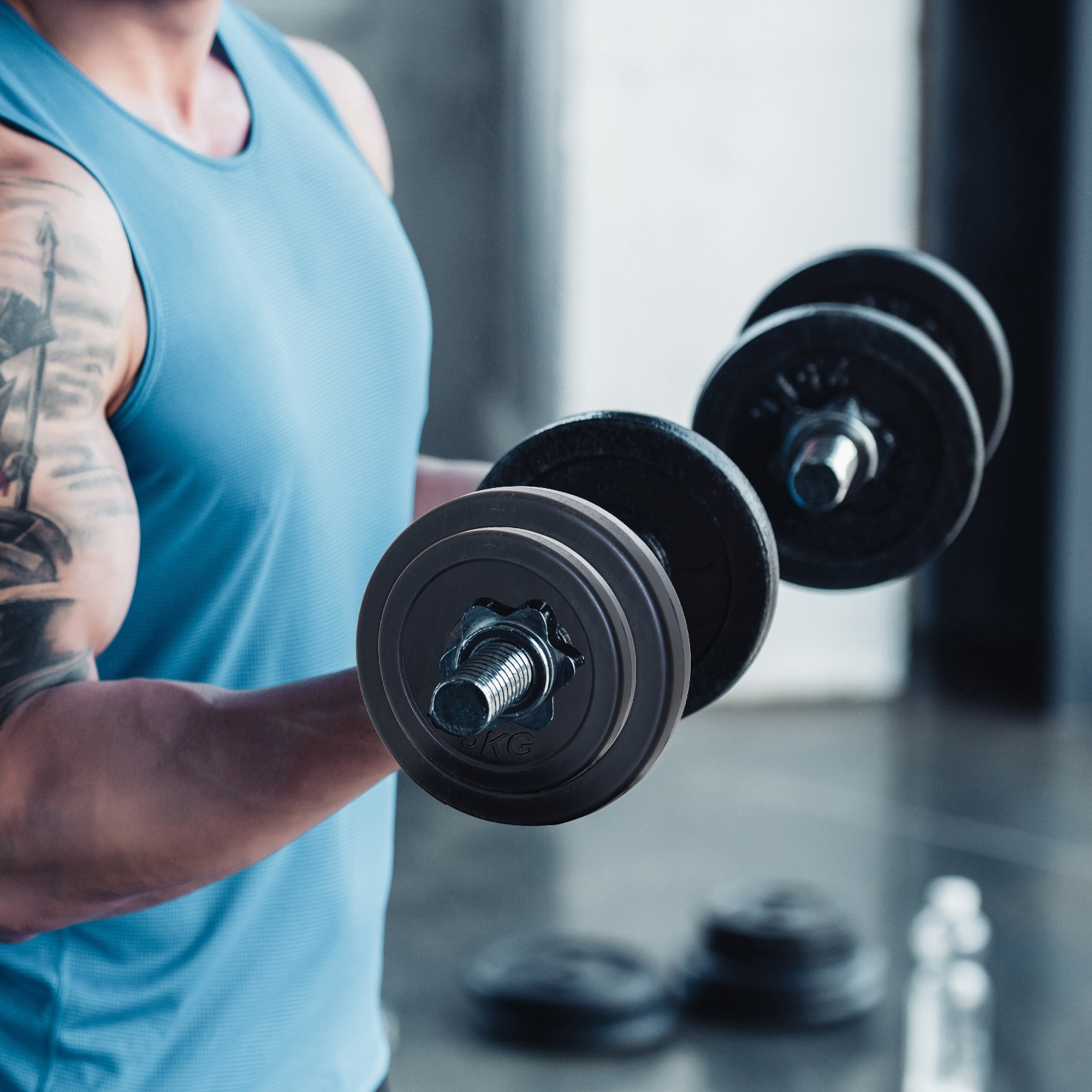 Un hombre con camiseta sin mangas azul hace curl con mancuernas. El gimnasio borroso al fondo.