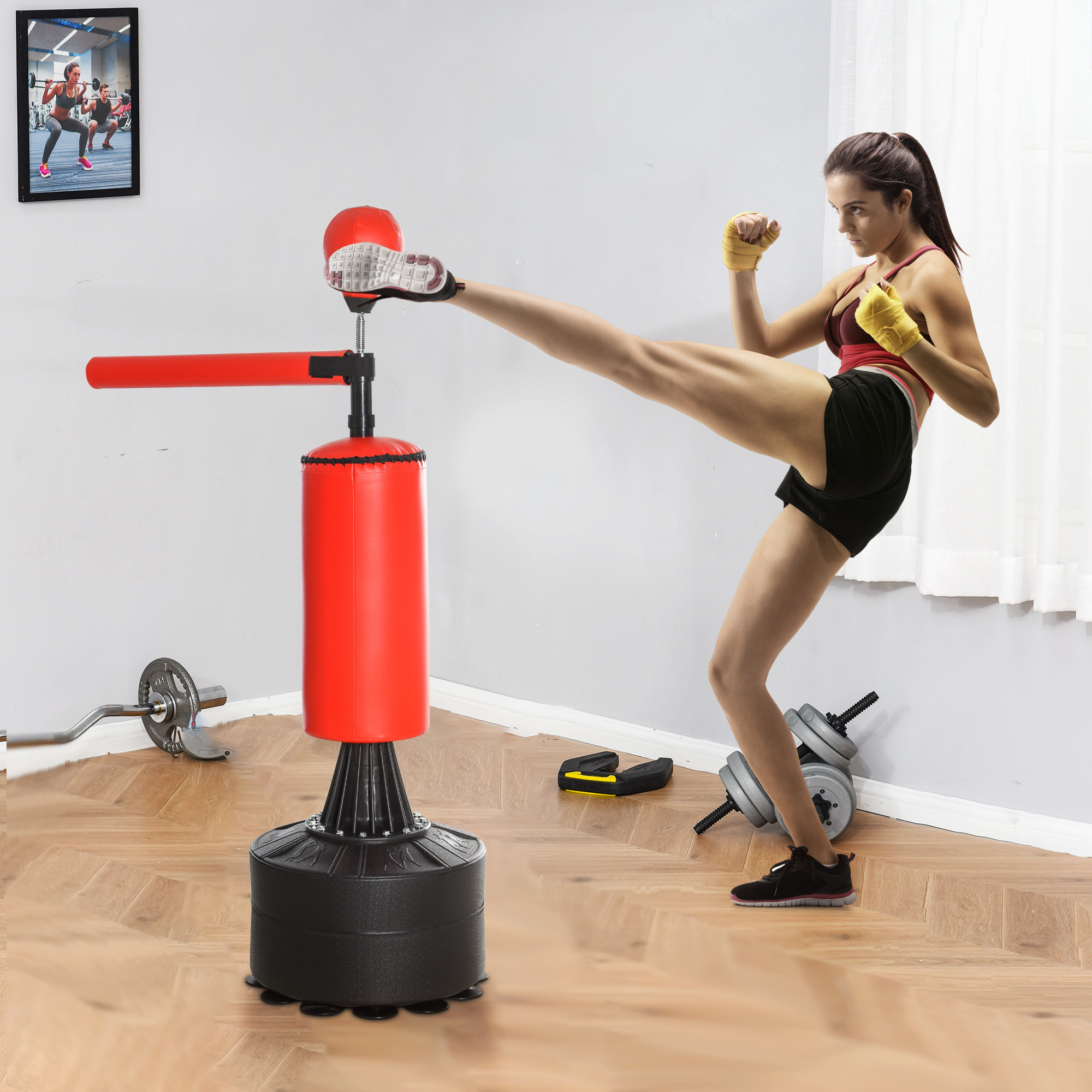 Una mujer patea un soporte de boxeo rojo y negro. Lleva pantalones cortos negros, una camiseta roja y vendas en las manos. Hay pesas cerca.