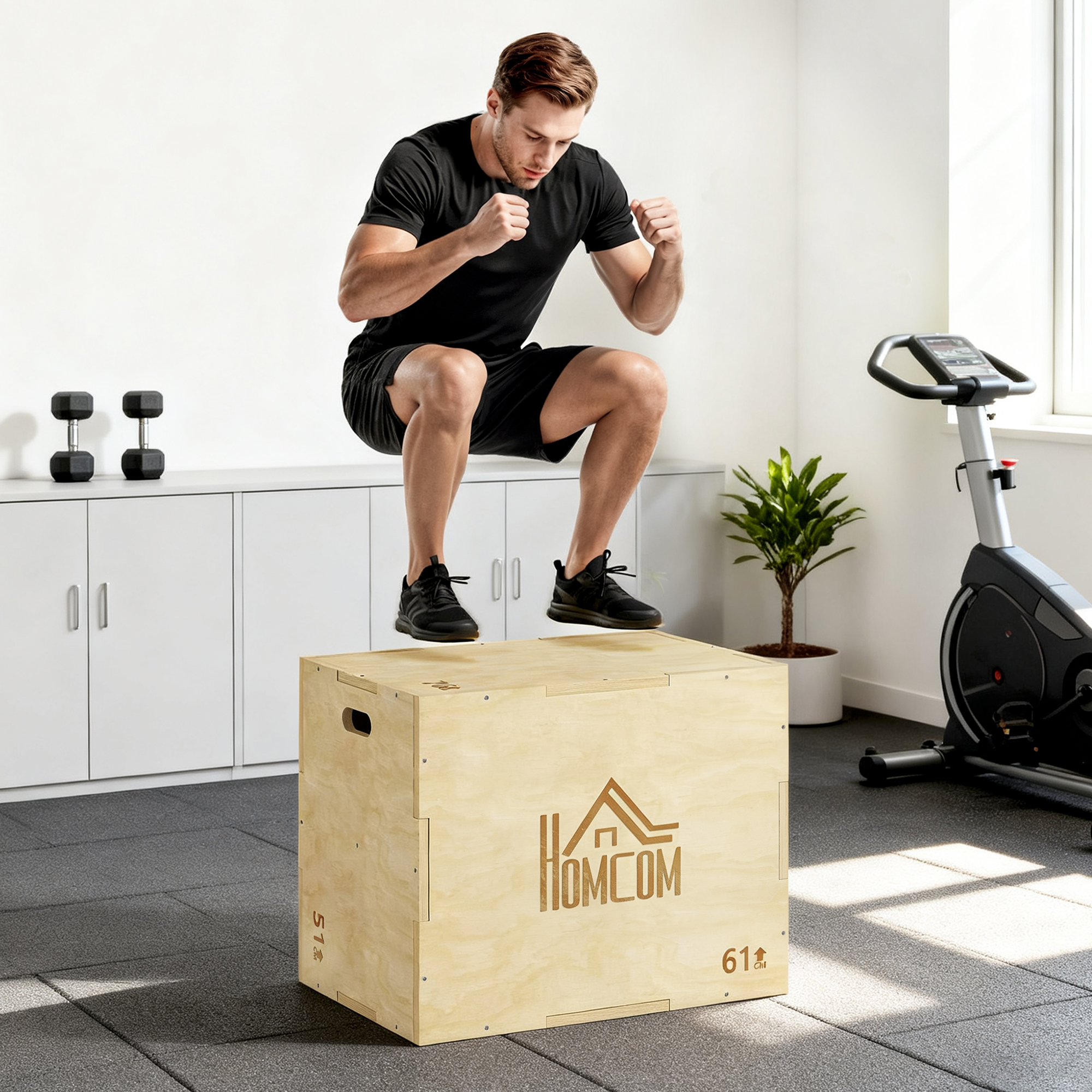 Un hombre salta sobre una caja de madera. Está en un gimnasio con pesas, una planta y una bicicleta estática al fondo.