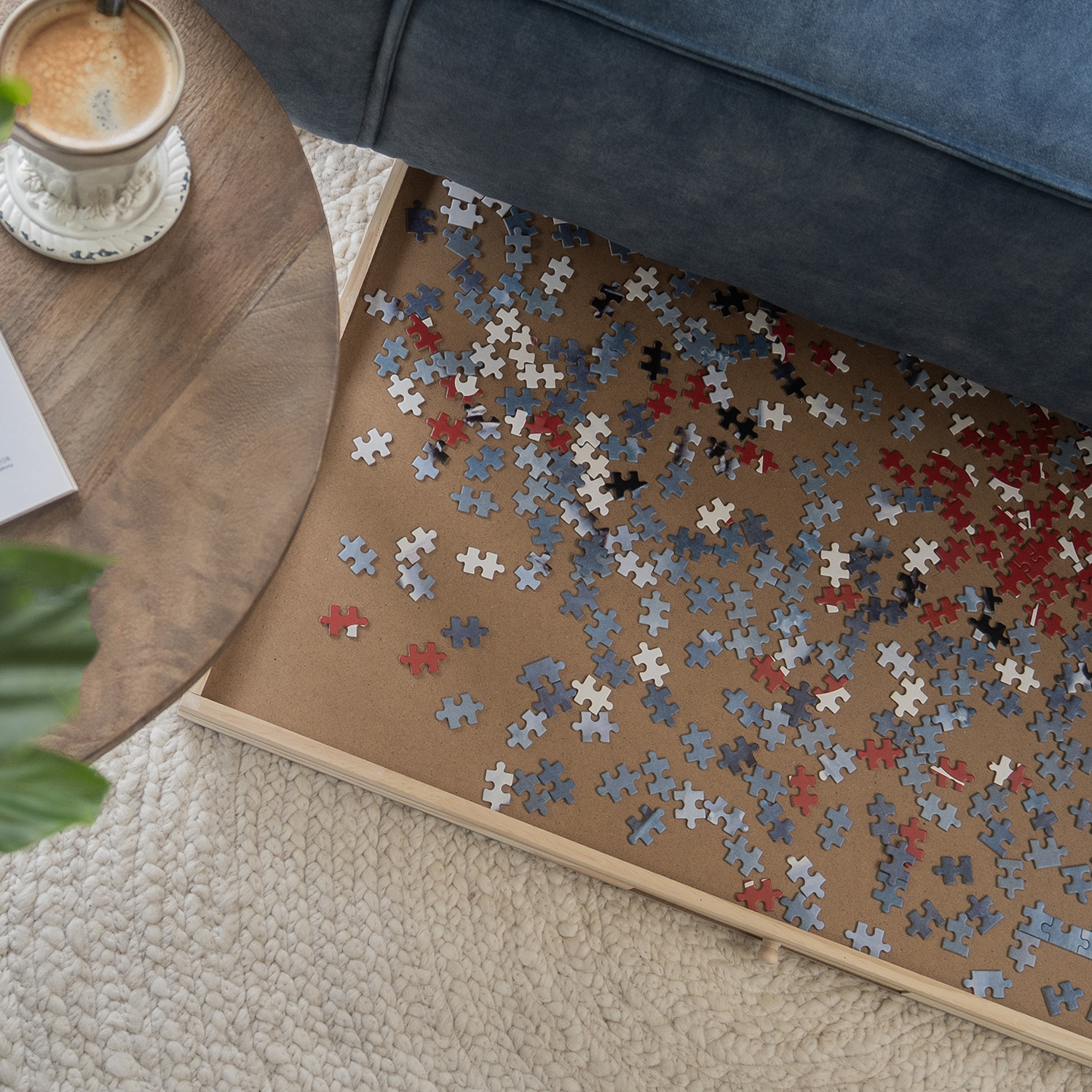 A coffee cup, a puzzle in progress, and a sofa are on a rug. Puzzle pieces are red, white, black, and blue. A table is nearby.