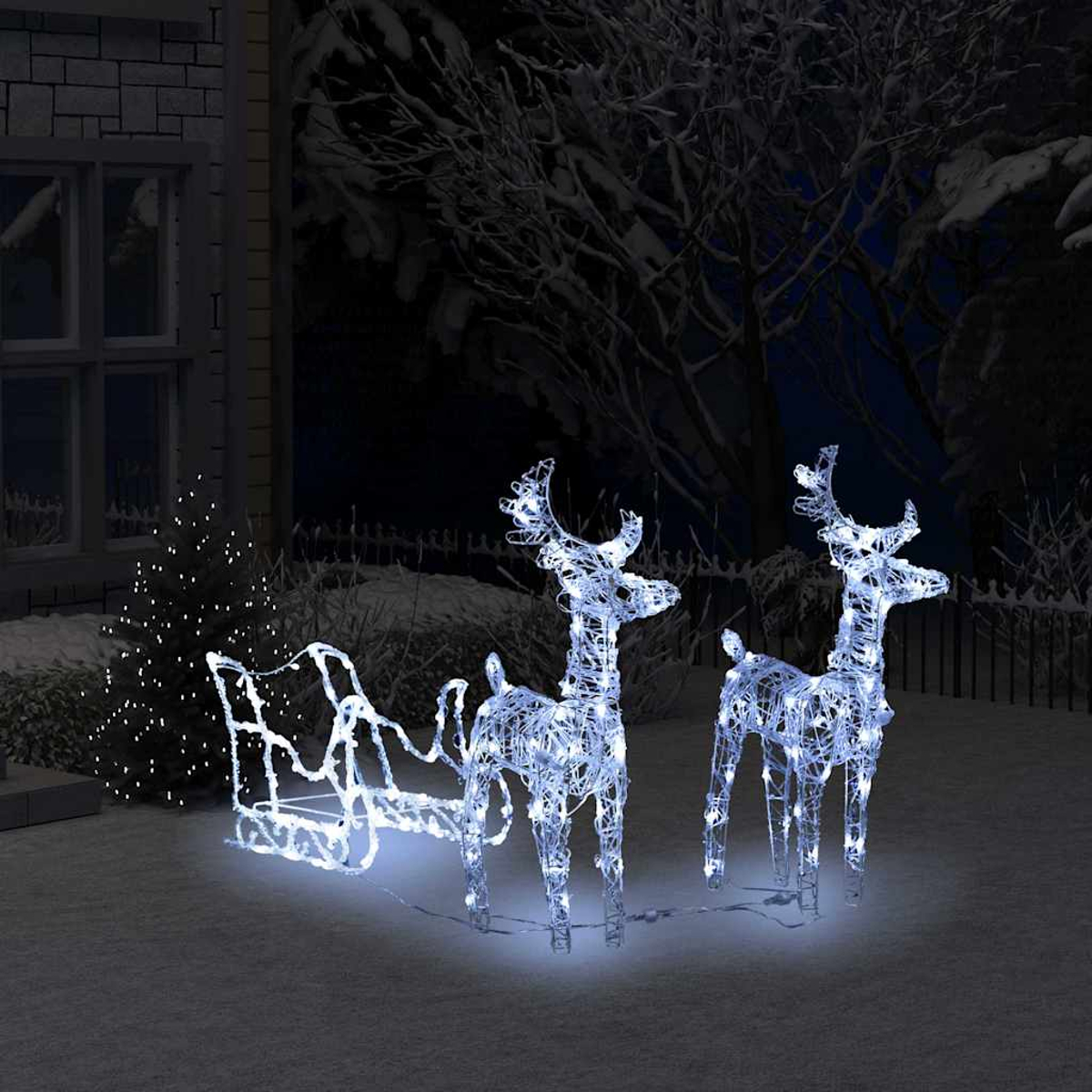 Beleuchtete Rentier- und Schlittendekorationen in einem verschneiten Garten bei Nacht. Ein beleuchteter Weihnachtsbaum und ein Haus sind im Hintergrund.