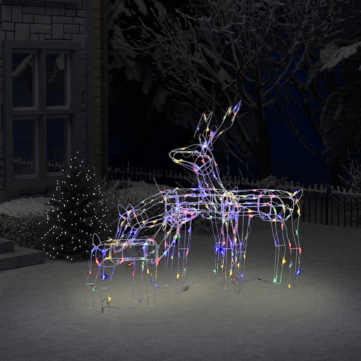 Zwei beleuchtete Drahtgestell-Rentiere mit bunten Lichtern stehen in einem verschneiten Garten. Ein Haus, ein Baum und der Nachthimmel sind im Hintergrund.