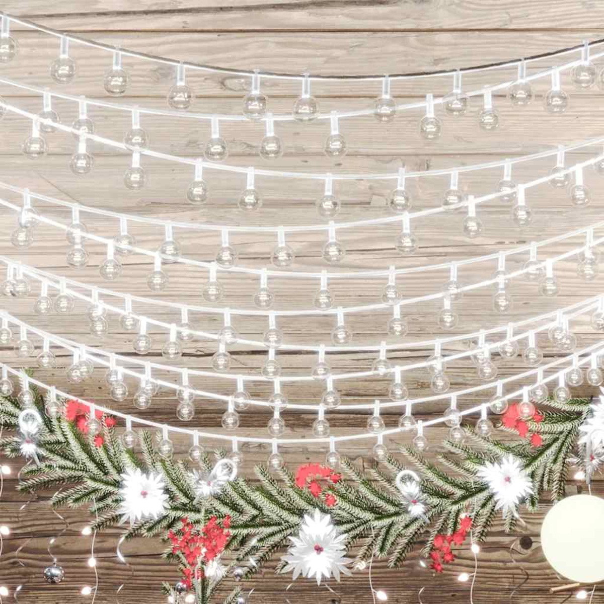 Lichterketten mit kleinen Glühbirnen hängen über einer Weihnachtsguirlande. Die Girlande hat Kiefernzweige, weiße Blumen und rote Beeren.