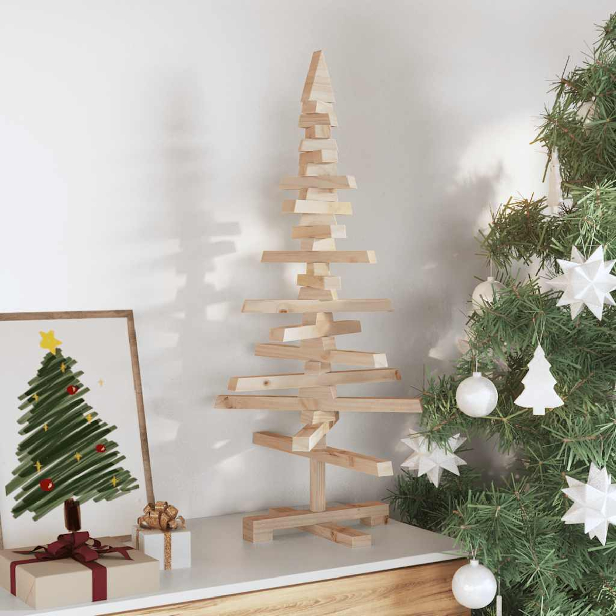Ein Weihnachtsbaum aus Holz und ein echter Baum stehen auf einer weißen Oberfläche. Eine gerahmte Zeichnung eines Baumes und Geschenke sind ebenfalls vorhanden.
