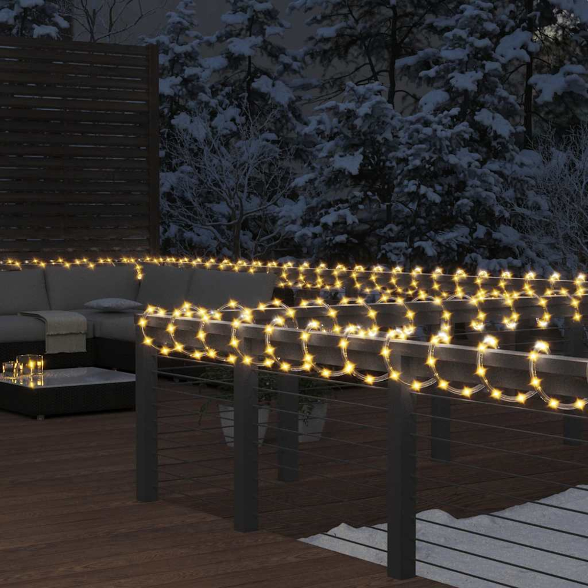Nachtszenen mit Lichterketten auf einem Deckgeländer. Im Hintergrund ein Sofa und verschneite Bäume.