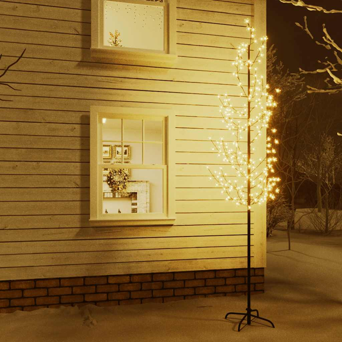 Ein beleuchteter Deko-Baum steht vor einem Haus mit Fenstern. Das Haus hat Holzverkleidung. Schnee bedeckt den Boden.