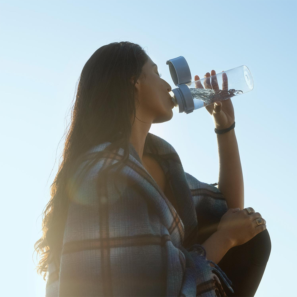 Kobieta pije wodę z przezroczystej butelki, owinięta w kratkowany koc na tle błękitnego nieba.
