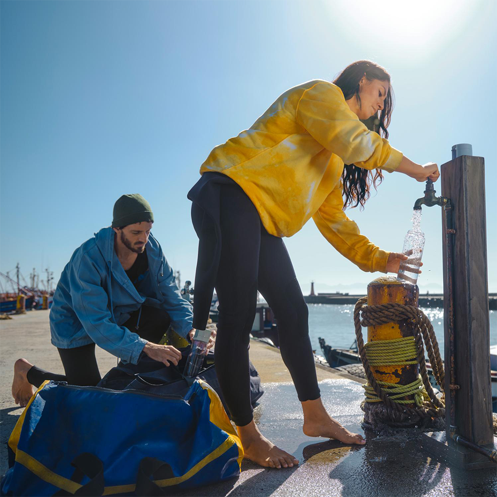 Kobieta napełnia butelkę wodą z kranu. Mężczyzna klęczy obok. Molo, łodzie i morze w tle.