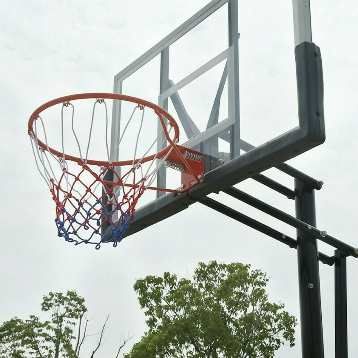Basketball hoop with clear backboard, orange rim, and net. Green trees are in the background under a cloudy sky.