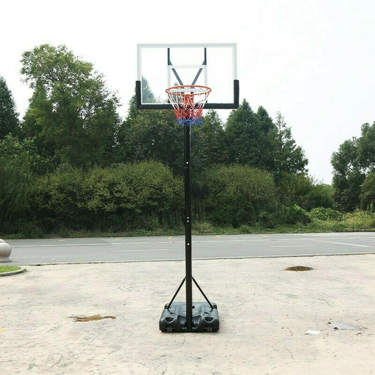 A basketball hoop on a black stand. The hoop is clear with a red and white net. Green trees are in the background.