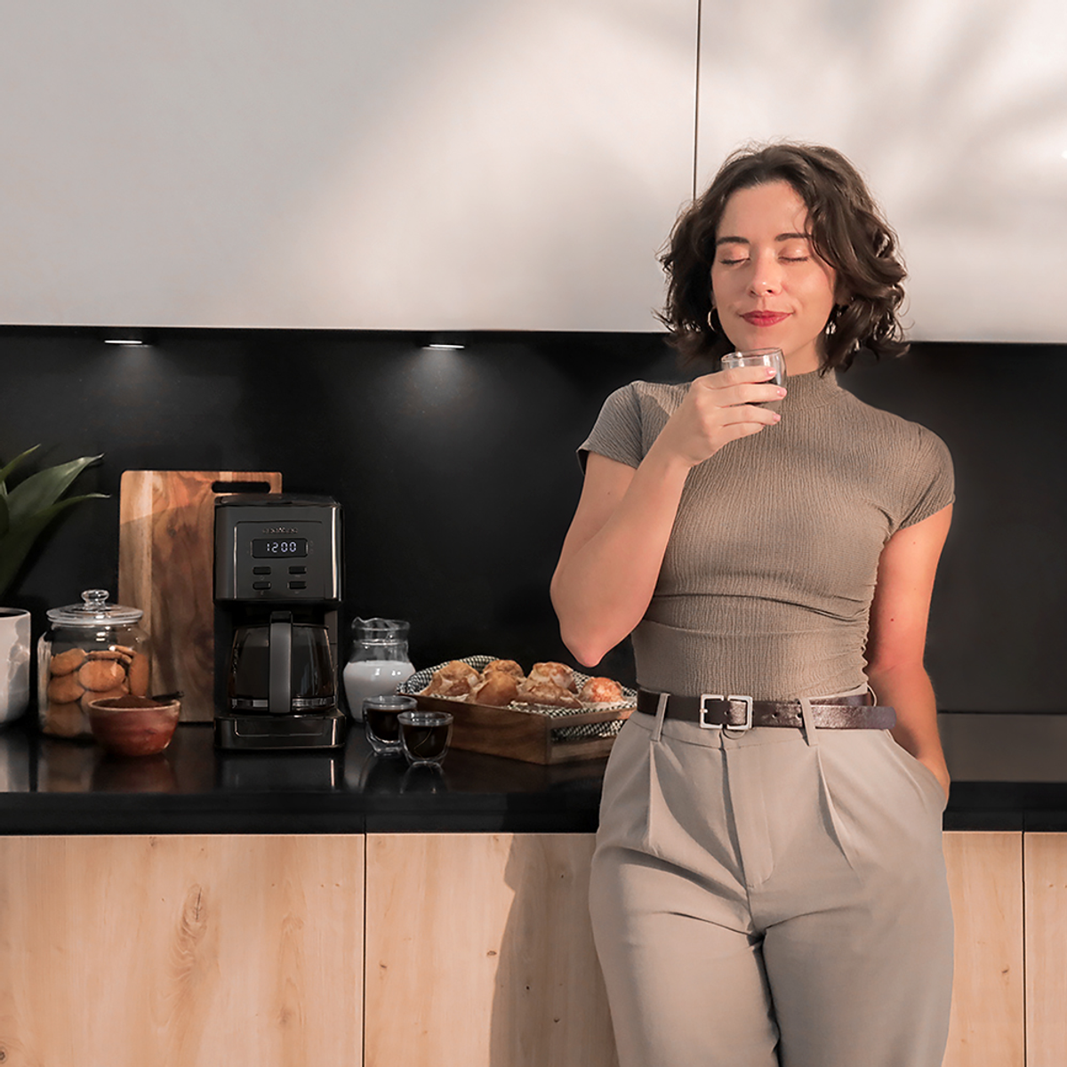 Kobieta w kuchni wącha kawę. Ekspres do kawy, ciasta i rośliny na blacie. Ma na sobie szary top i spodnie.