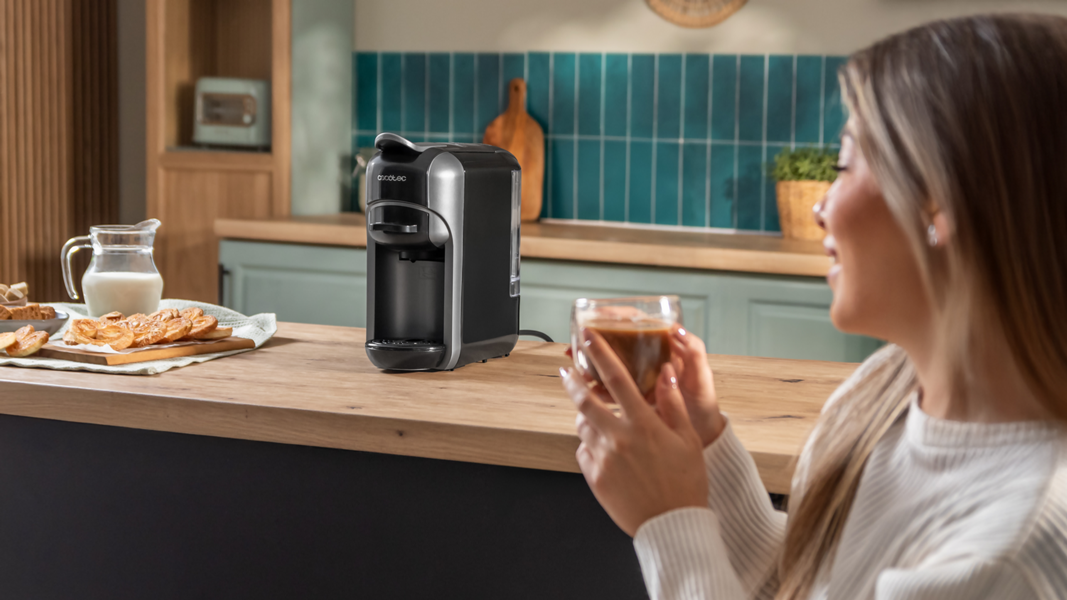 Eine Frau hält Kaffee in der Nähe einer Kaffeemaschine. Im Hintergrund sind Küchentheke mit Gebäck, Milch und eine türkisfarbene Fliesenwand zu sehen.
