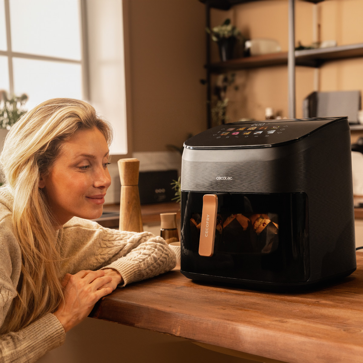 Kobieta patrzy na czarną frytkownicę na drewnianym blacie. Frytkownica wyświetla jedzenie. Tło pokazuje okno i półki.