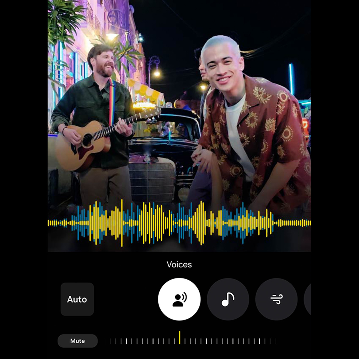 Dwóch mężczyzn na ulicy. Jeden gra na gitarze, drugi się uśmiecha. Stary samochód w tle.