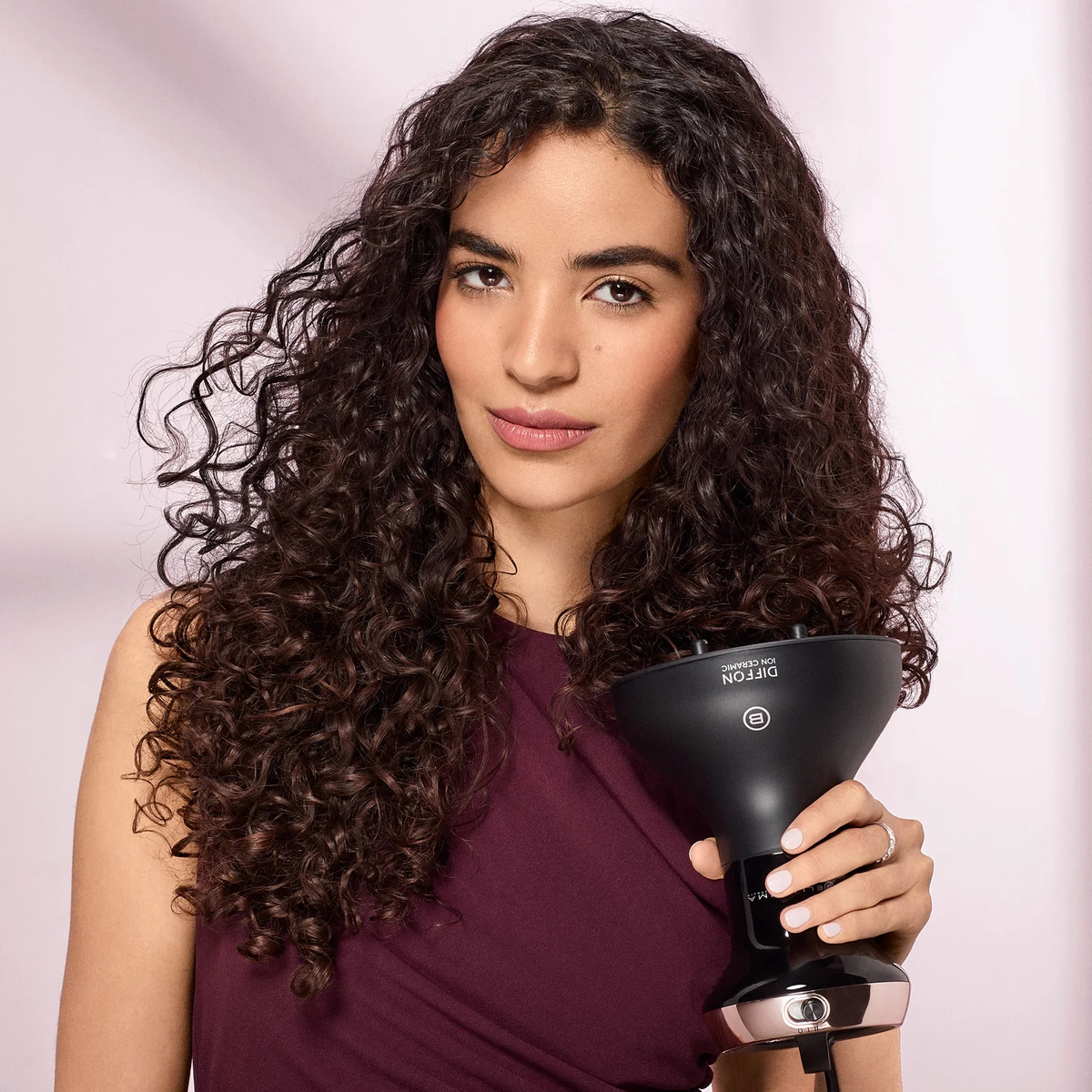 Kobieta z kręconymi włosami trzyma czarny dyfuzor do włosów. Ma na sobie bordową sukienkę. Tło jest jasnoróżowe.