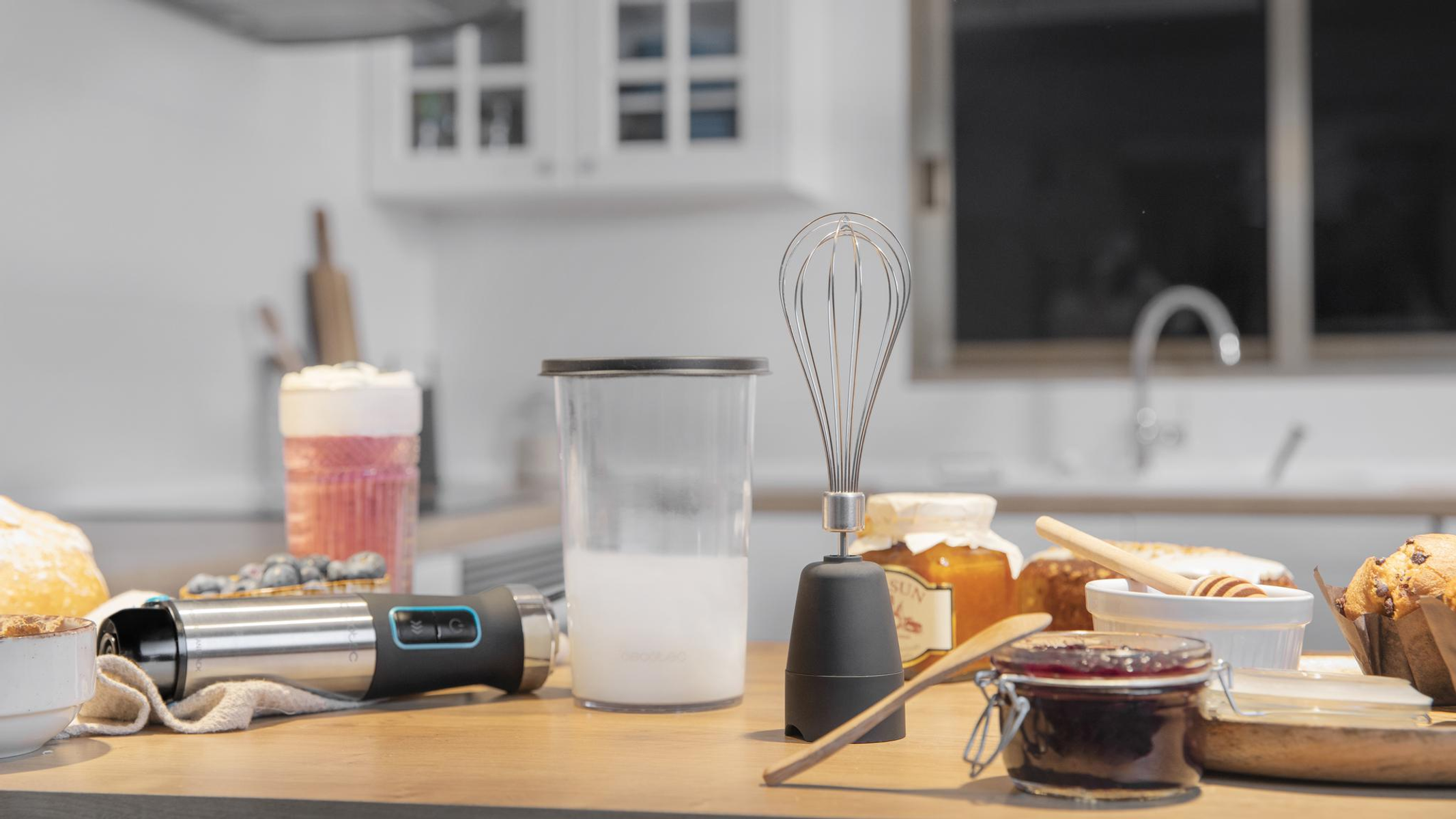 Un piano cucina con frullatore, frusta, barattolo di marmellata, miele, pane e un bicchiere con liquido rosa.