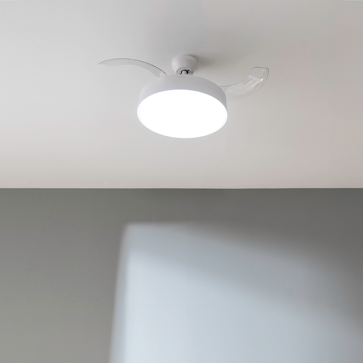 Un ventilador de techo blanco con luz circular y dos aspas está montado en un techo blanco. Una pared gris es visible debajo.