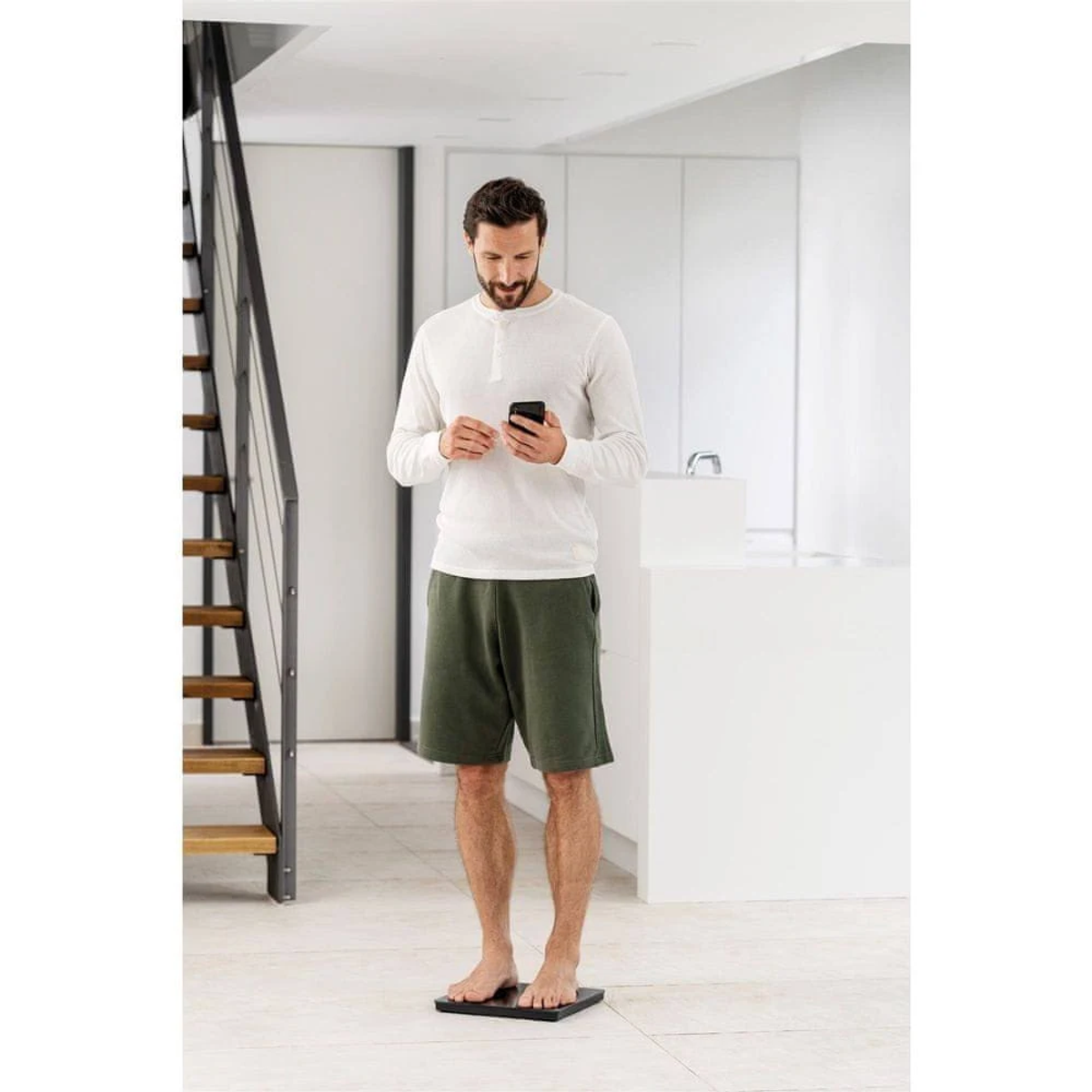 Ein Mann in weißem Hemd und grünen Shorts steht auf einer Waage und schaut auf sein Handy. Eine Treppe und weiße Schränke sind im Hintergrund.
