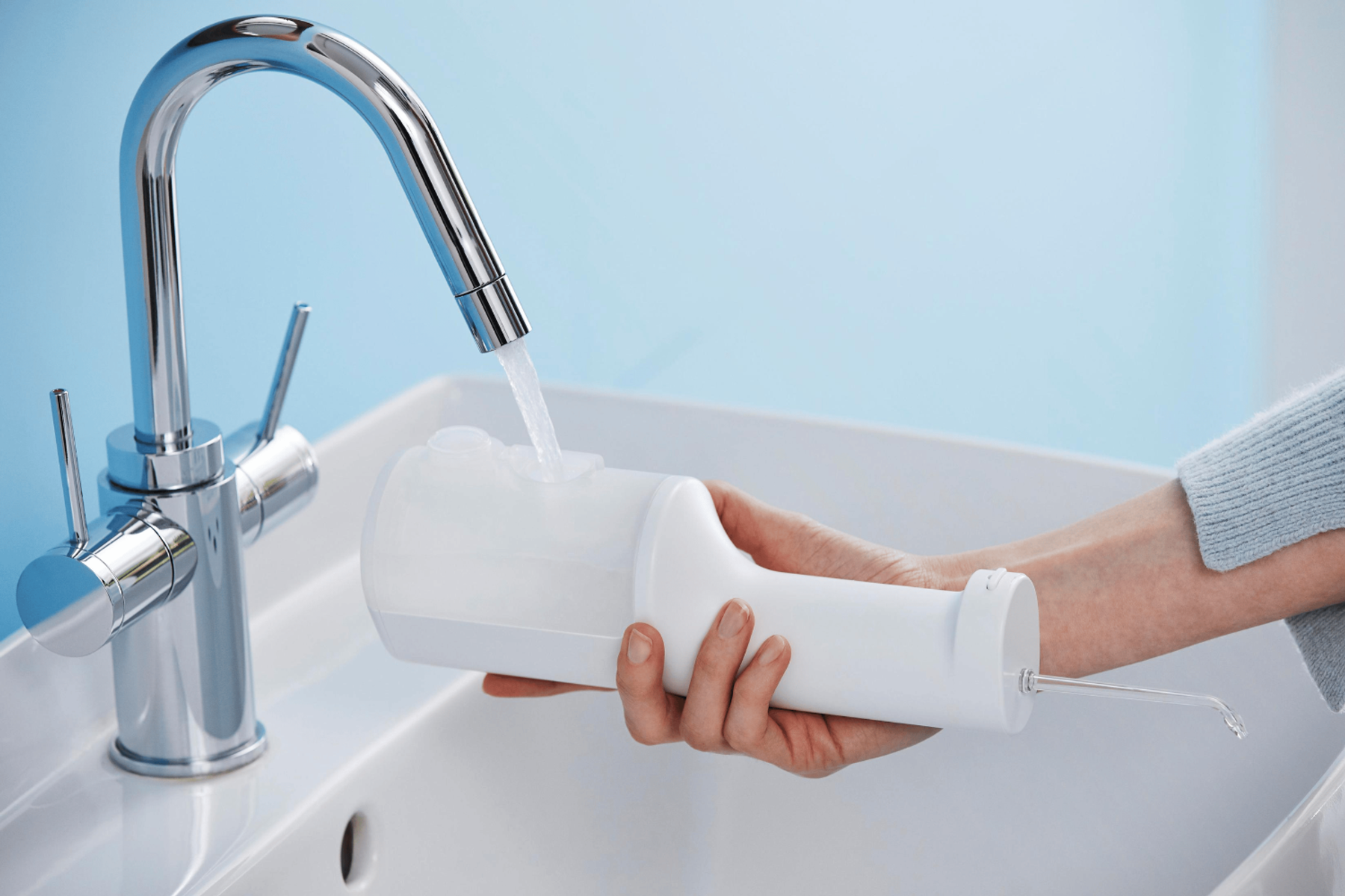 Eine Person hält einen weißen Munddusche unter einem Wasserhahn mit fließendem Wasser. Der Hintergrund ist eine blaue Wand und ein weißes Waschbecken.