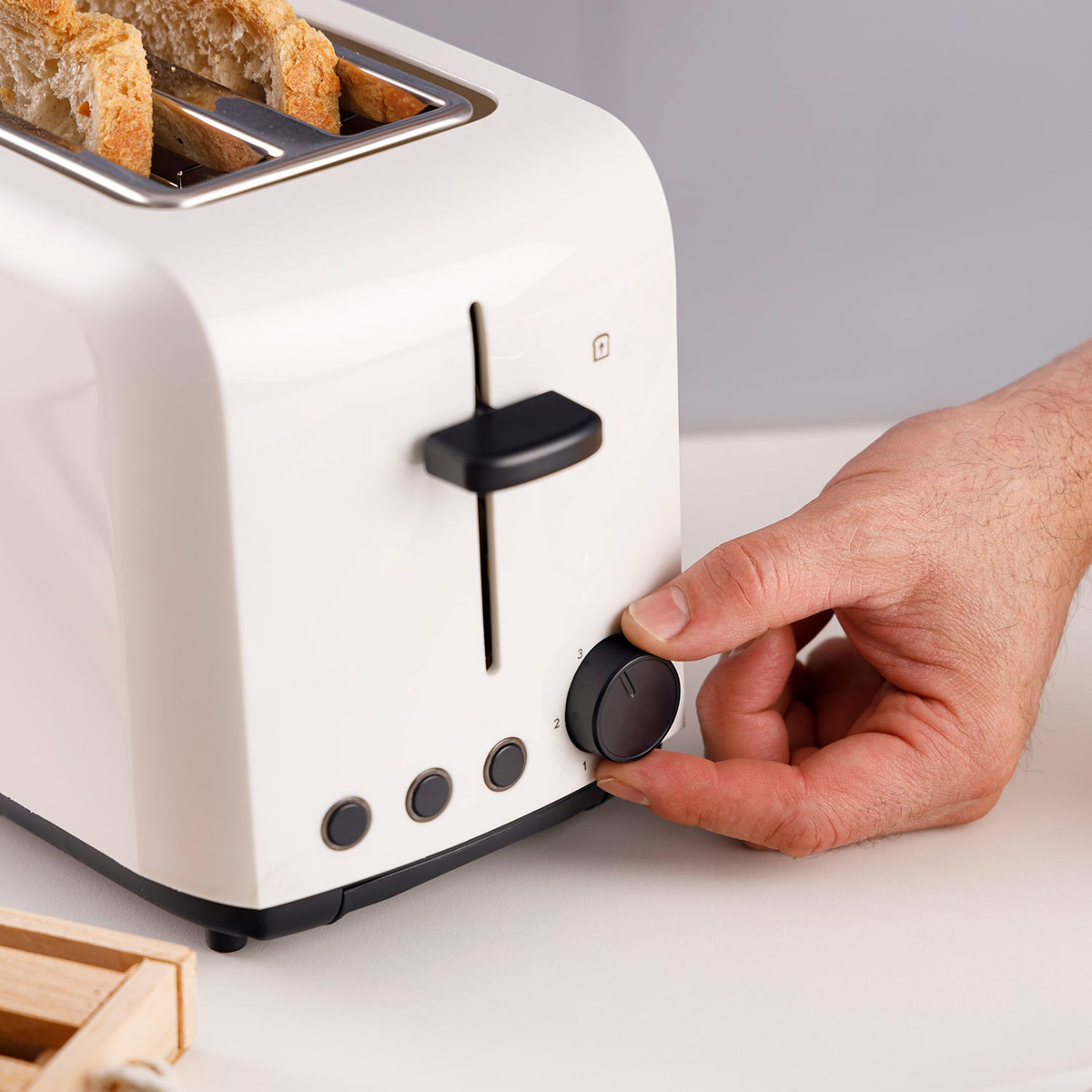 Biały toster z kromkami chleba w środku. Ręka obraca pokrętło regulacji zarumienienia. Widoczna drewniana skrzynka.