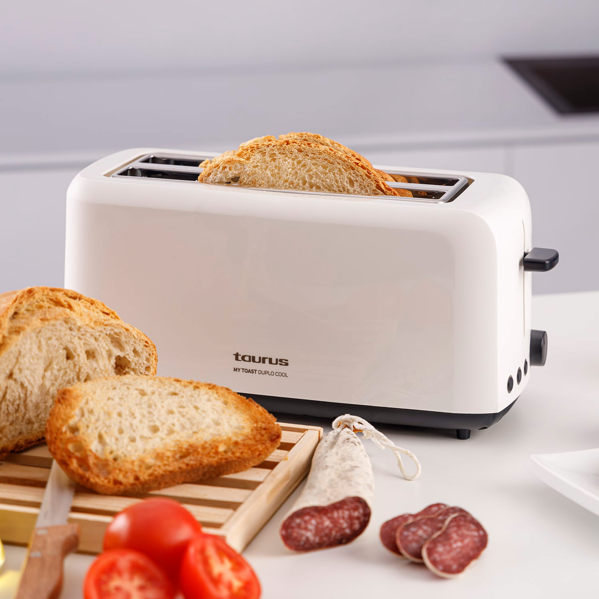 Biały toster z chlebem w środku. Na stole leżą kromki chleba, pomidory, kiełbasa i nóż.