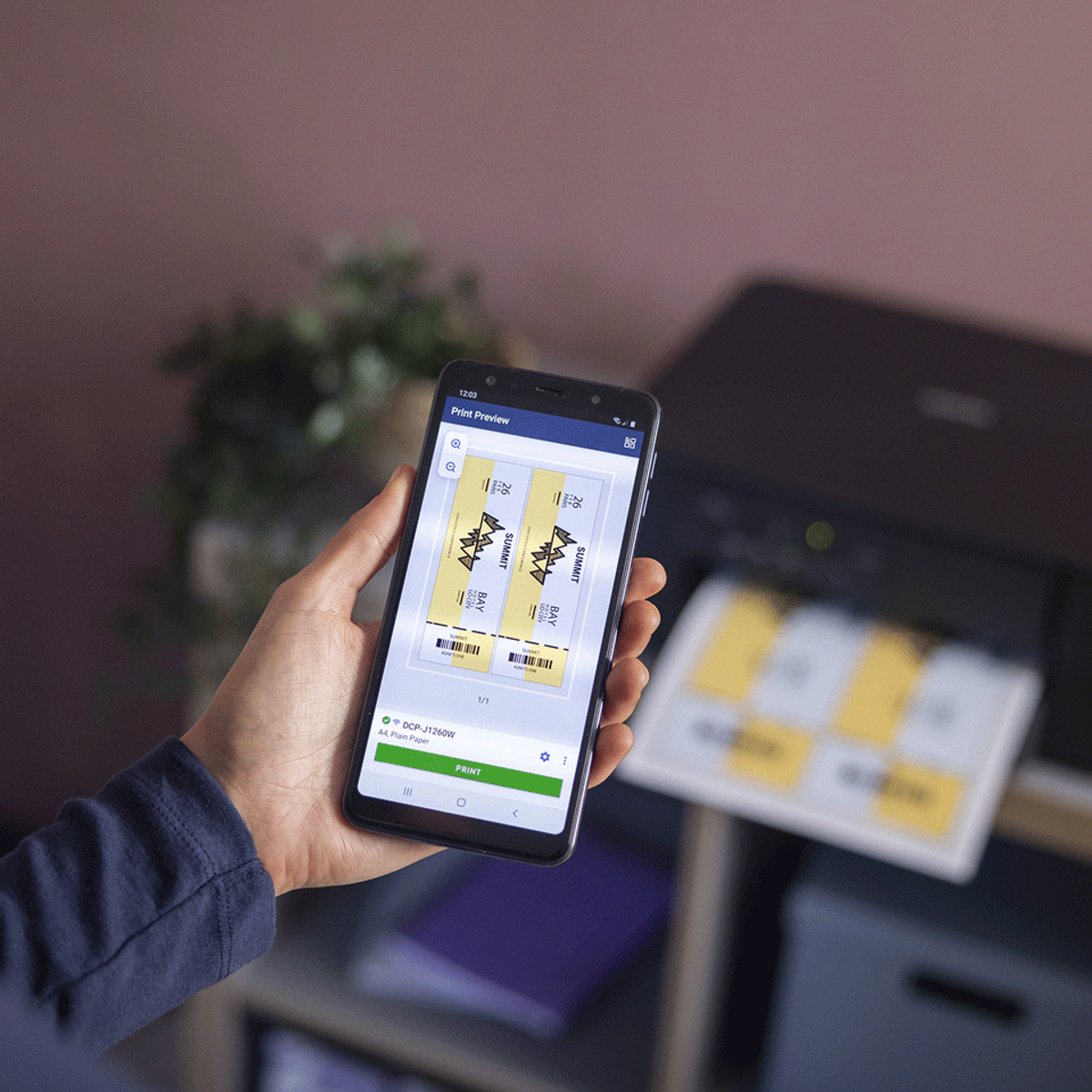 Ręka trzymająca telefon z podglądem wydruku biletów. W tle drukarka, widoczne wydrukowane bilety.