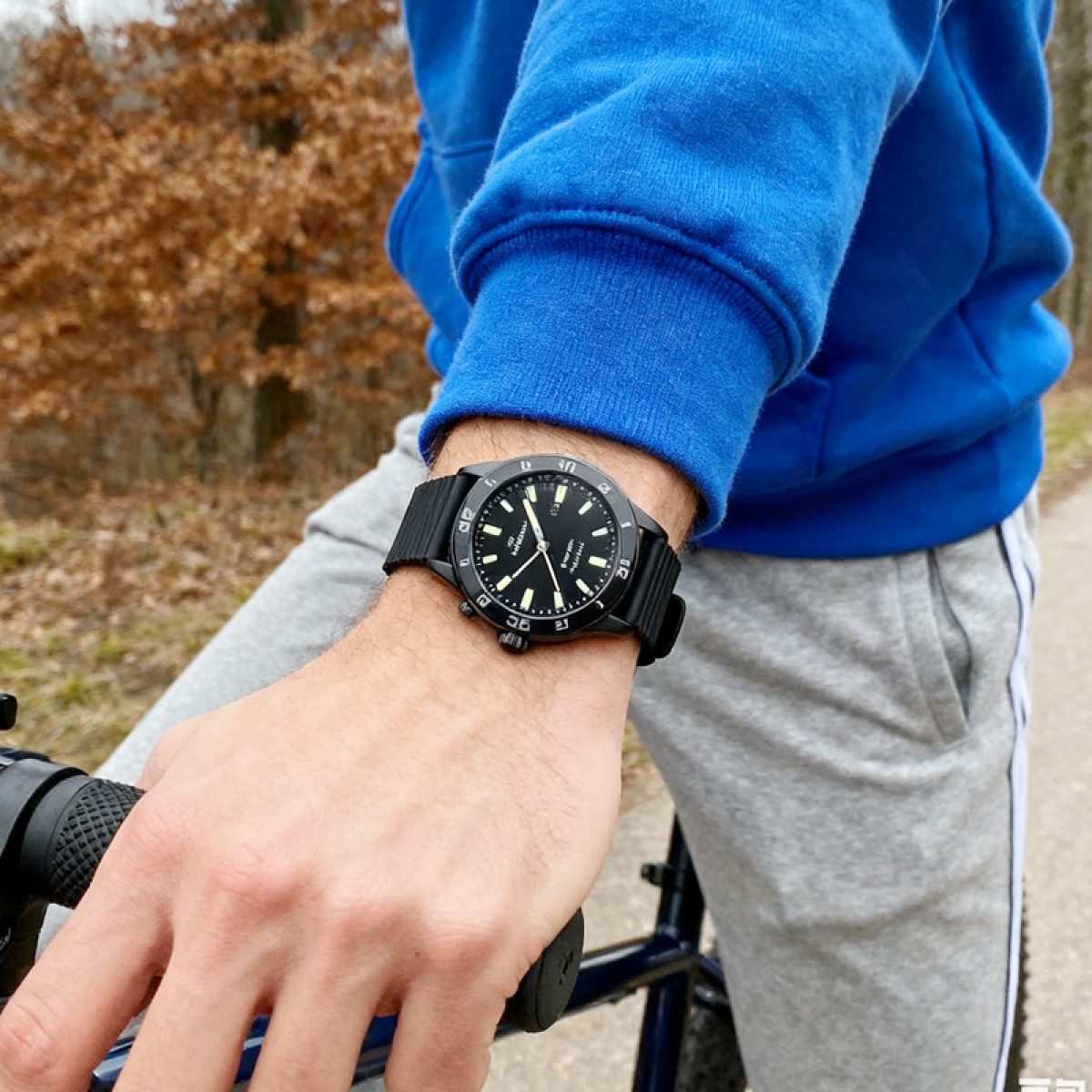 Osoba nosząca czarny zegarek, niebieską bluzę z kapturem i szare spodnie dresowe trzyma rower. Tło to las z brązowymi liśćmi.