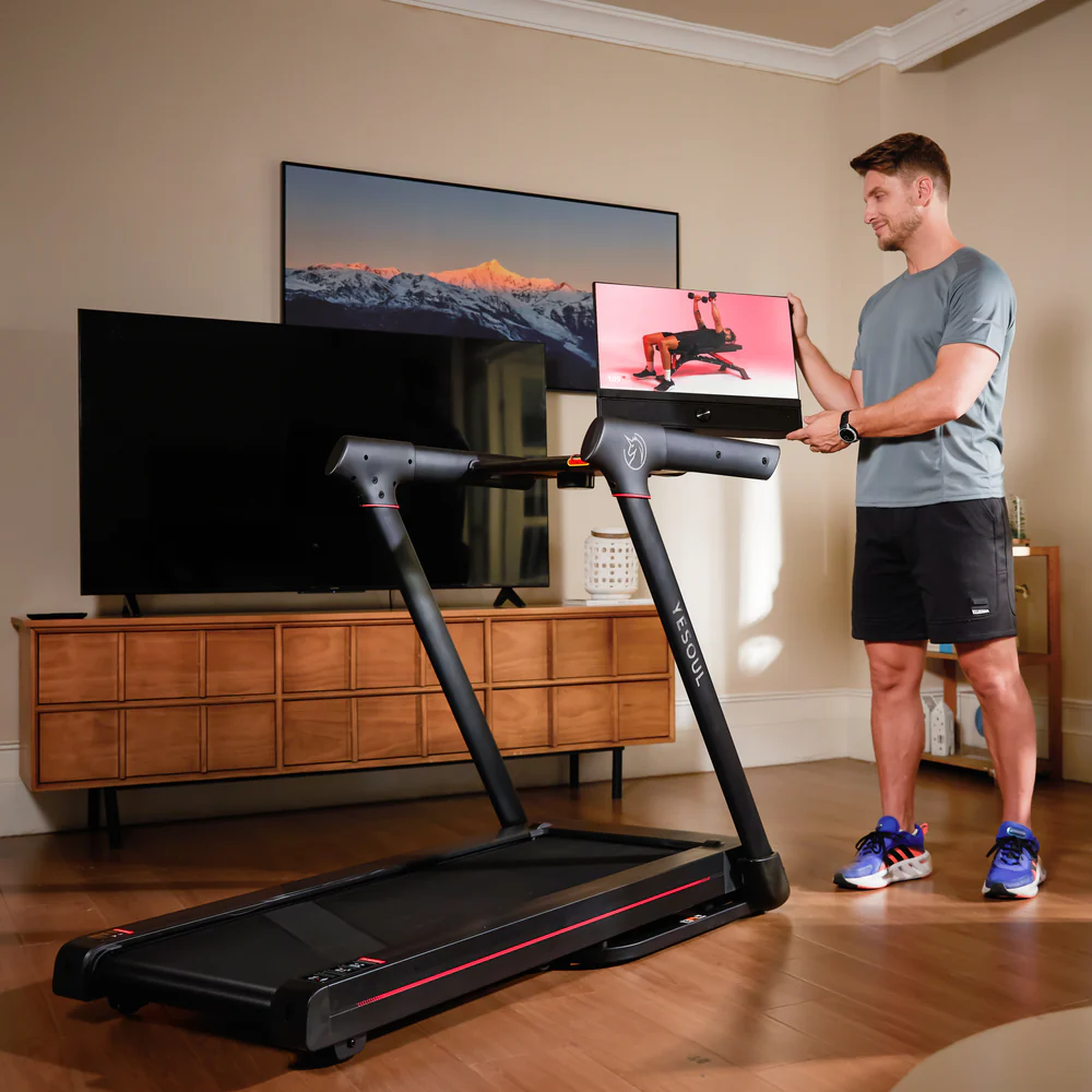 Hombre de pie con pantalla mostrando video de entrenamiento, cerca de una cinta de correr. El fondo incluye un televisor, un mueble de madera y una imagen de montaña.