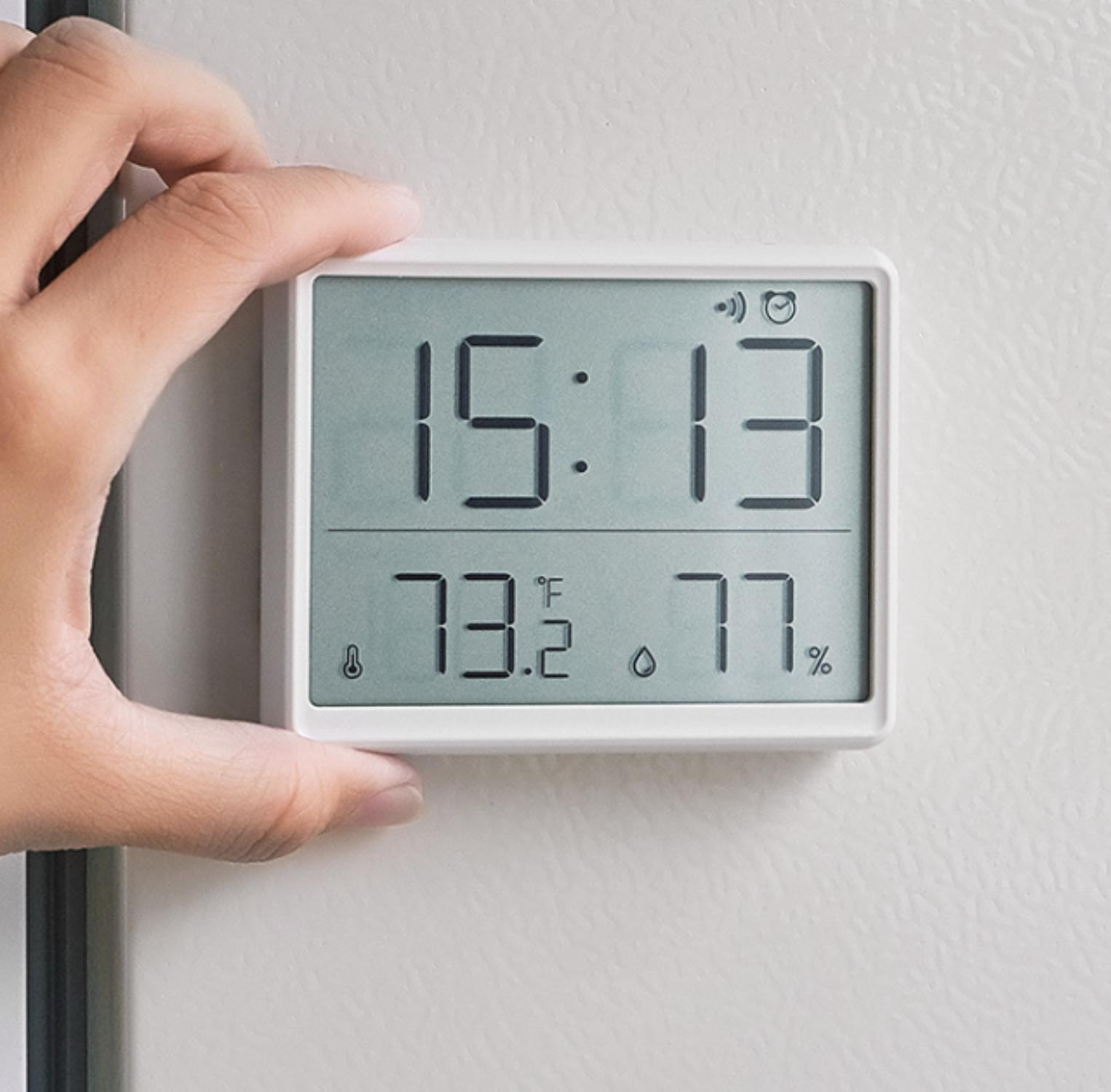 Eine Hand hält ein weißes digitales Thermometer, das 15:13, 73,2 °F und 77 % anzeigt. Der Hintergrund ist eine weiße Oberfläche.
