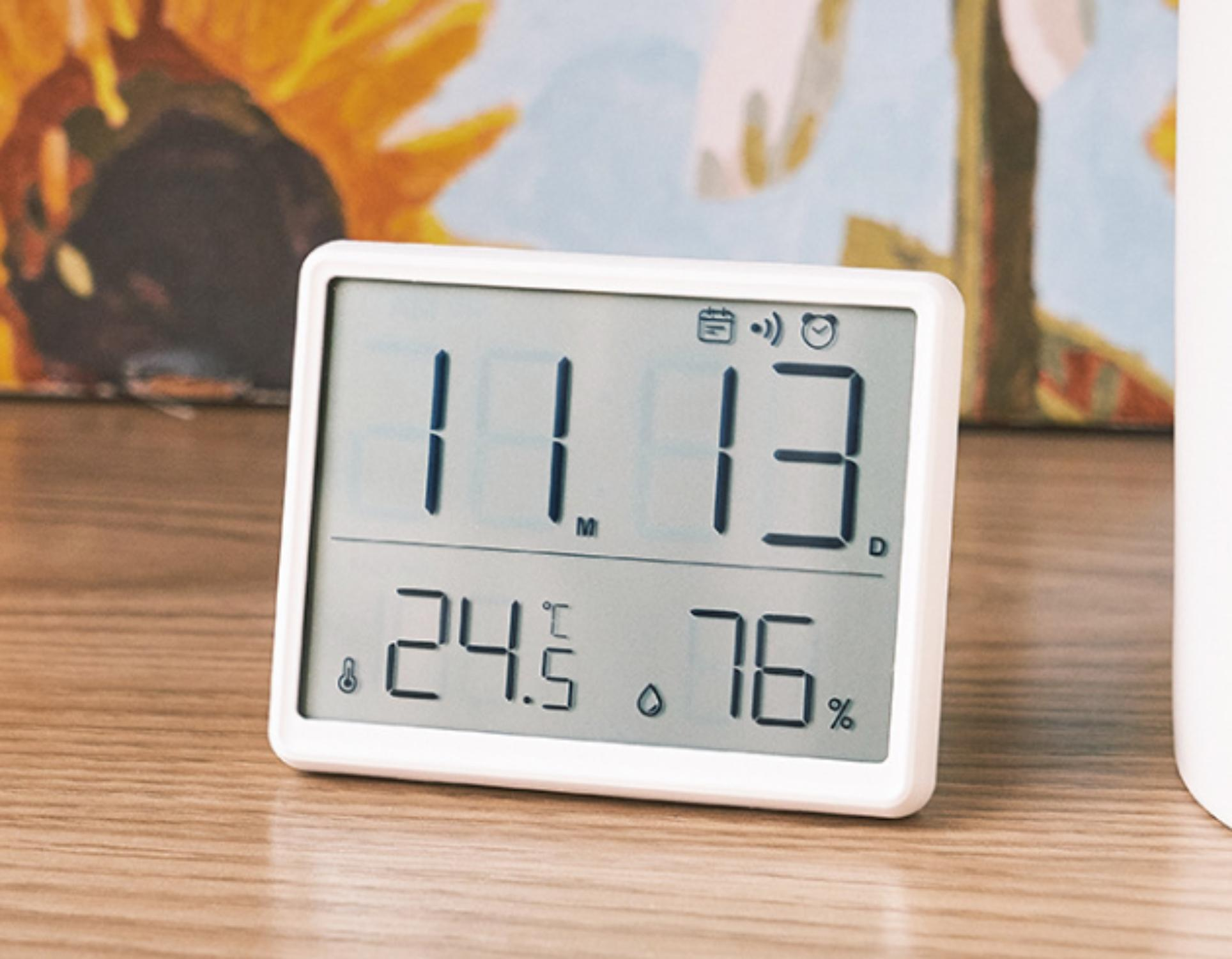Eine weiße Digitaluhr zeigt Datum 11.13, Temp. 24,5°C und Luftfeuchtigkeit 76% auf einem Holztisch, mit einem bunten Gemälde im Hintergrund.