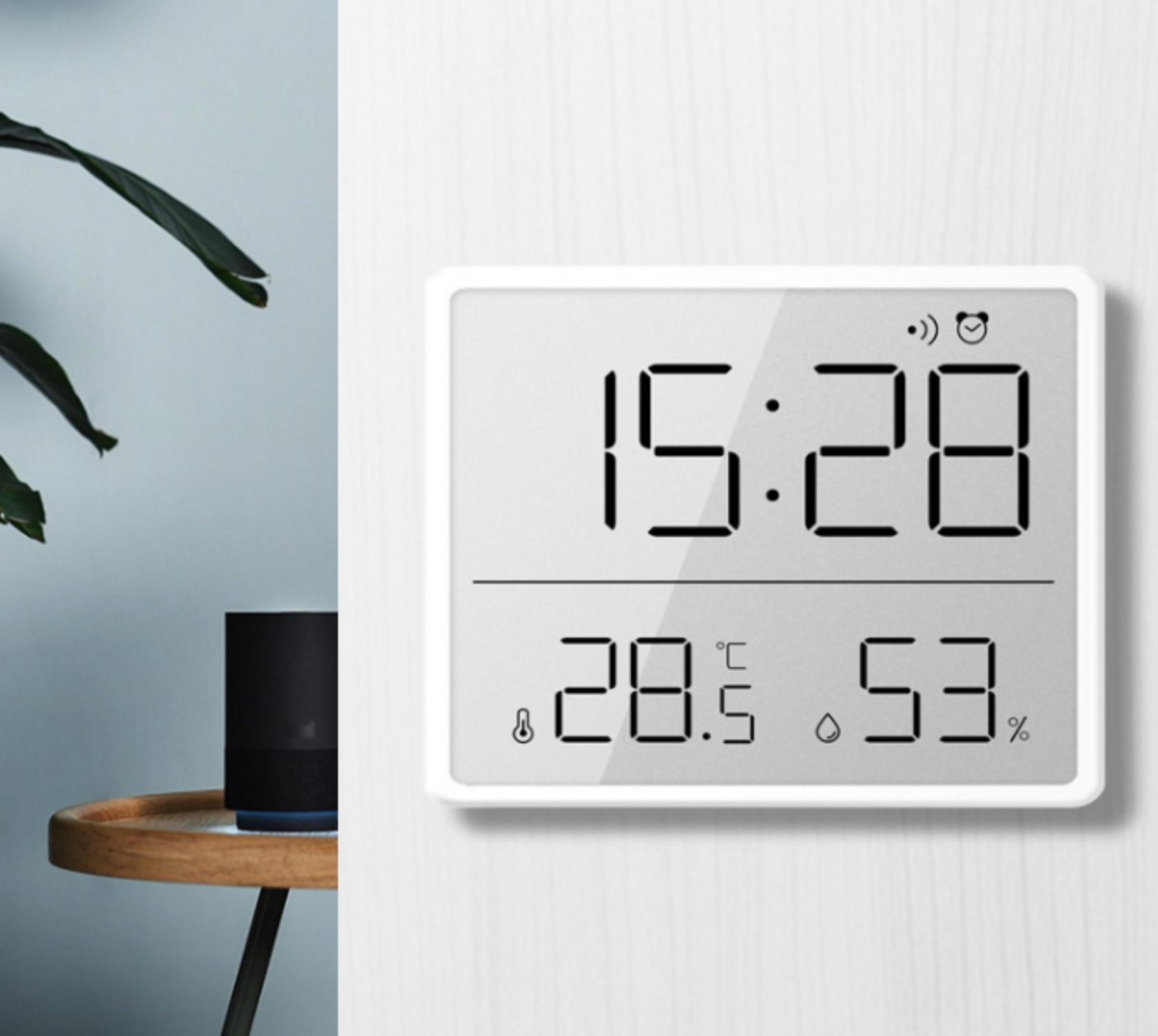 Eine Digitaluhr zeigt 15:28, 28,5 Grad Celsius und 53 % Luftfeuchtigkeit an. Ein schwarzer Lautsprecher steht auf einem Holztisch.