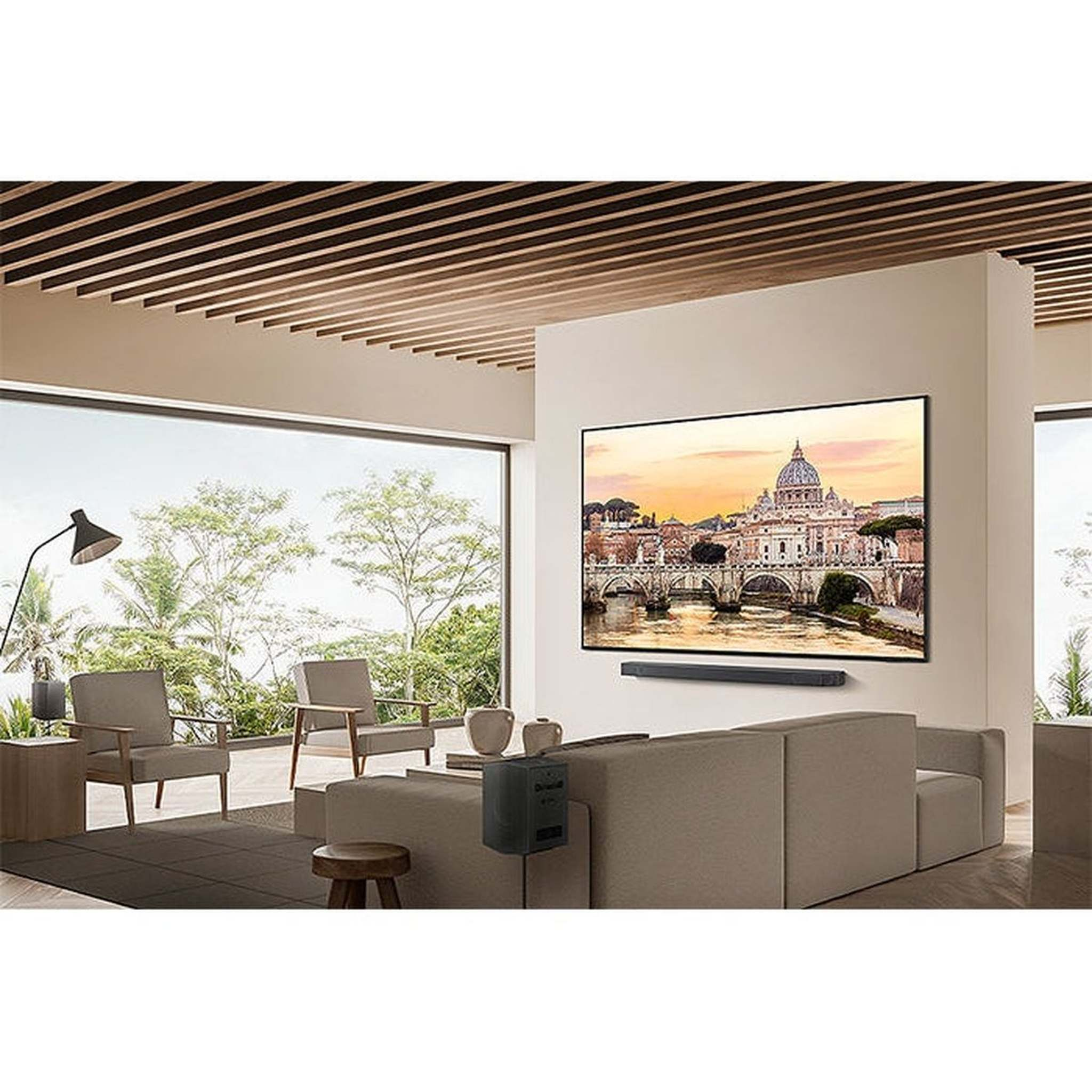 Soggiorno con grande TV che mostra una scena cittadina, divano, sedie e grandi finestre con vista su alberi e cielo.