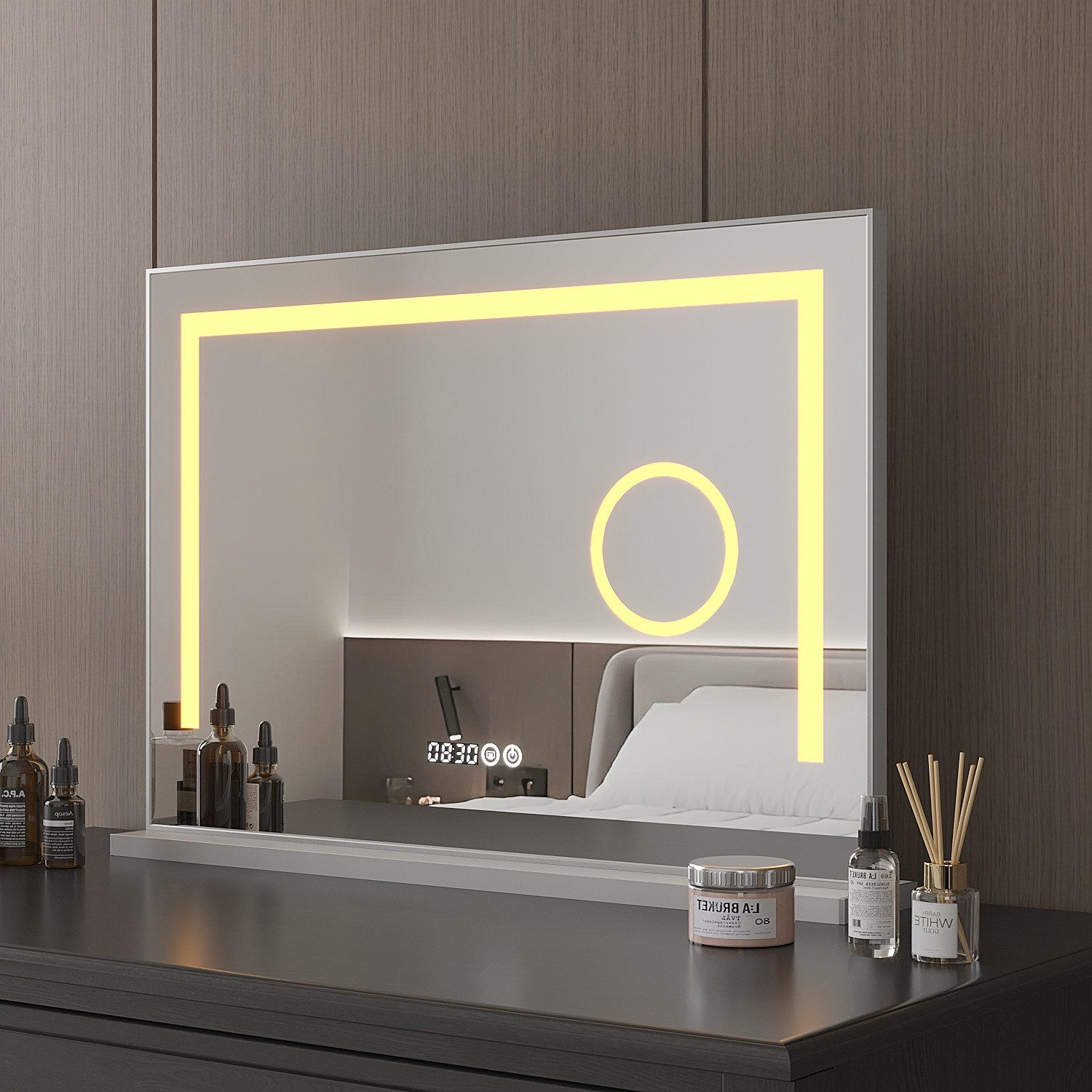 Ein beleuchteter Spiegel auf einer dunklen Kommode spiegelt ein Schlafzimmer wider. Flaschen und ein Glas stehen auf der Kommode. Ein Bett spiegelt sich im Spiegel.